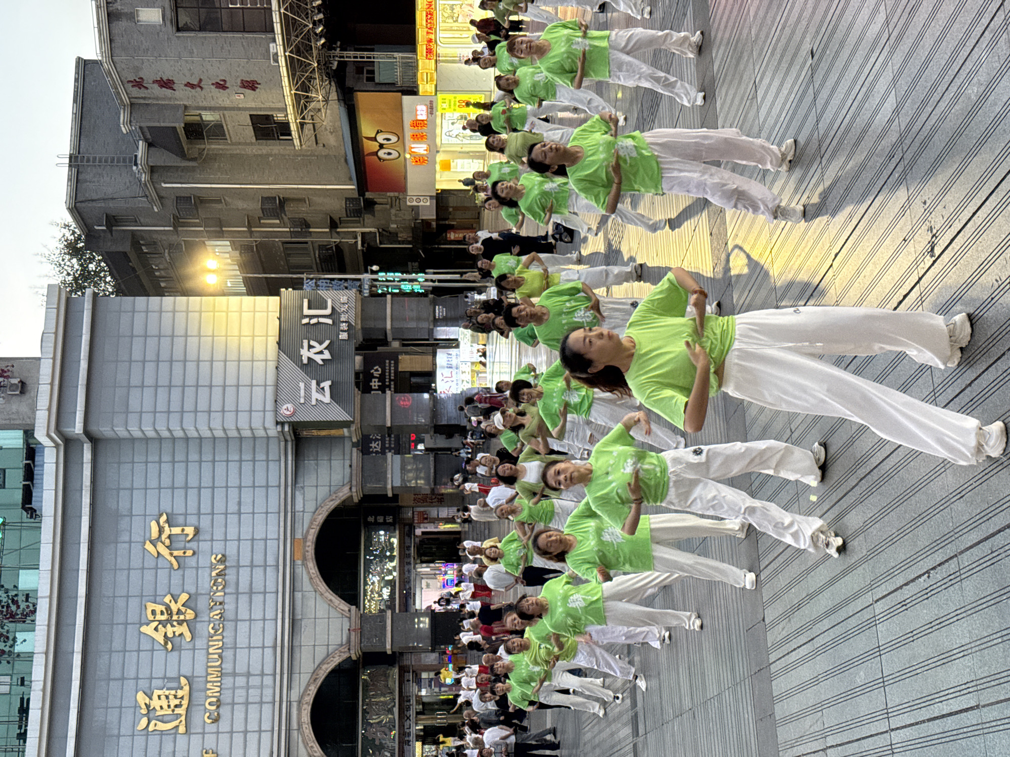 This outdoor photograph captures a group of women in Beibei, China, engaged in a synchronized exercise routine. The women, all wearing lime green shirts and white pants, are arranged in rows.

The backdrop includes urban architecture, with a building on the left featuring the Chinese characters 通銀行 COMMUNICATIONS, rendered in gold, and signage indicating businesses such as 北店, 云衣汇 服装批发, and 达. Additional signage, including CHOW TAISENG, is visible.

The pavement is marked with linear patterns. Spectators are observing in the background. The photograph appears to have been taken at ground level.