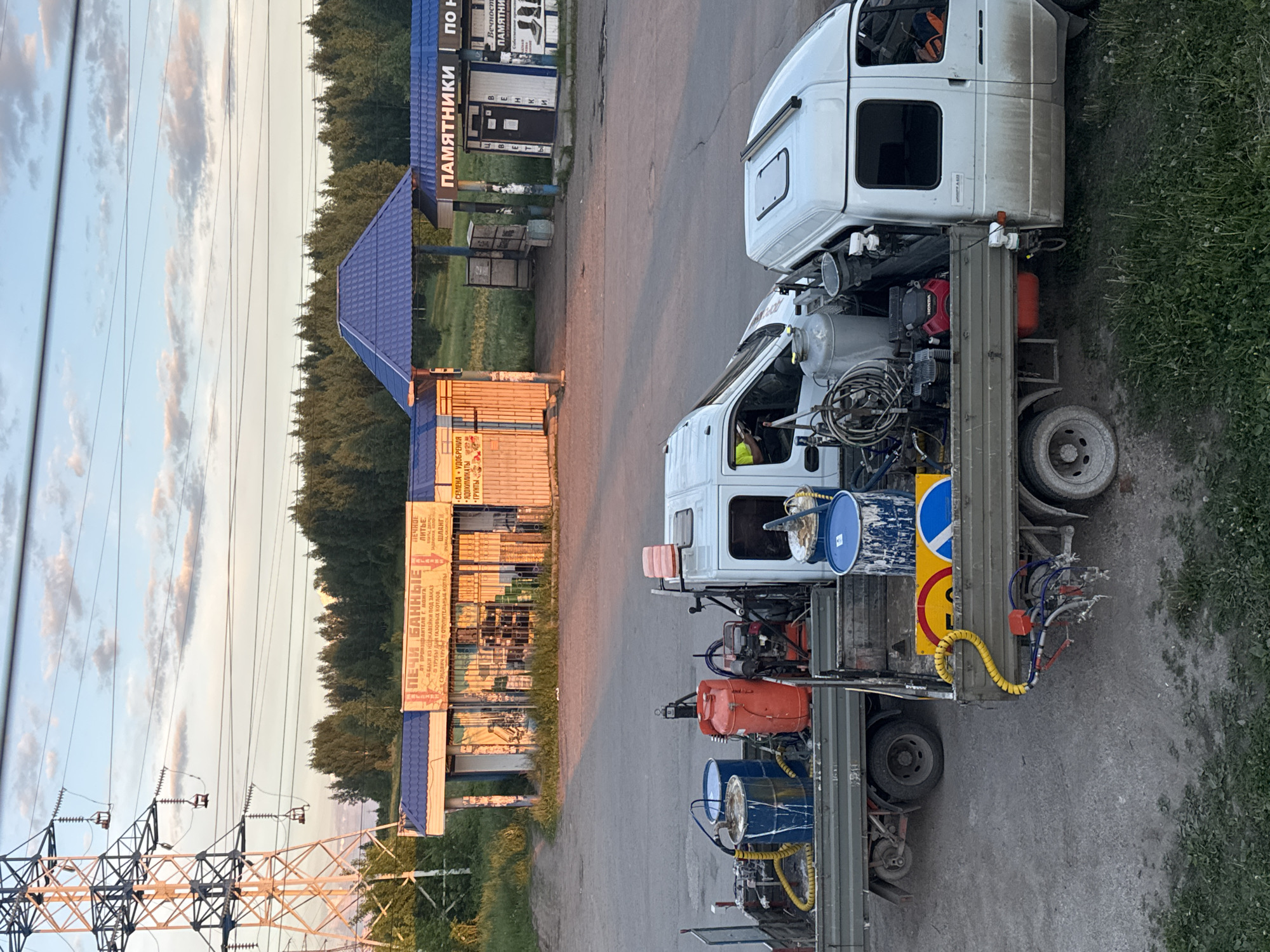 The image depicts an outdoor scene in Mozhga, Russia, during late afternoon or early evening, characterized by soft, warm light and scattered clouds in a light blue sky. The setting appears to be a semi-industrial or commercial area alongside a paved road, bordered by grassy patches and dense trees in the background.
Two white commercial trucks are prominent in the foreground and midground. The truck closer to the viewer is outfitted with specialized equipment, including large blue and orange barrels or tanks, a red Honda engine, and a network of hoses and nozzles, strongly suggesting it is a road marking vehicle. Road signs, including a yellow diamond-shaped sign with a right-turn arrow and a blue circular mandatory right-turn sign, are also visible on its flatbed. The other truck, similar in model, is parked behind it.
In the background, several buildings feature blue roofs. A main building with yellow-orange walls displays a large sign advertising "ПЕЧИ БАННЫЕ" (BATH OVENS) and "ОТ ПРОИЗВОДИТЕЛЯ Г. МОЖГА" (FROM THE MANUFACTURER G. MOZHA), confirming the location. Other visible text on the sign includes "БАКИ ИЗ НЕРЖАВЕЙКИ ПОД ЗАКАЗ" (STAINLESS STEEL TANKS TO ORDER), "ПЕЧНОЕ ЛИТЬЕ" (OVEN CASTING), and "ШЛАНГИ" (HOSES). An adjacent sign advertises "СЕМЕНА • УДОБРЕНИЯ" (SEEDS • FERTILIZERS), "ЯДОХИМИКАТЫ" (PESTICIDES), and "ГРУНТЫ" (SOILS). A taller sign on a pillar reads "ПАМЯТНИКИ" (MONUMENTS) and "Вечност" (Eternity), indicating a business selling monuments.
To the left, a large power pylon and overhead power lines traverse the sky. No individuals are visible, but the specialized equipment on the trucks implies ongoing or imminent work related to road infrastructure. The overall impression is of a functional, commercial setting at dusk.