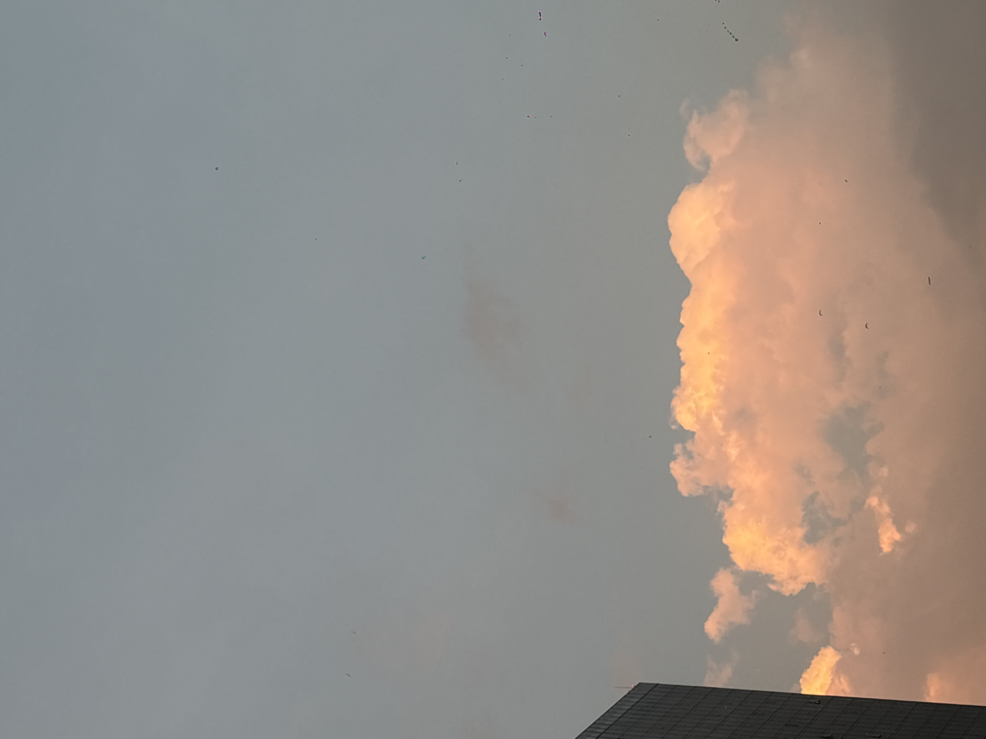 The image depicts a partly cloudy sky with a building visible at the bottom. The sky is a pale grey, with a large, fluffy cloud formation dominating the right side of the frame. The cloud appears to be illuminated by the setting sun, giving it a warm, orange hue. Small specks, possibly birds, are scattered across the sky. A section of a tall building, likely a commercial or residential structure in Mexico City, Mexico, is visible in the lower right corner. The building appears to be multi-storied with a dark, grid-like facade. The scene is peaceful, possibly during dusk or dawn.
