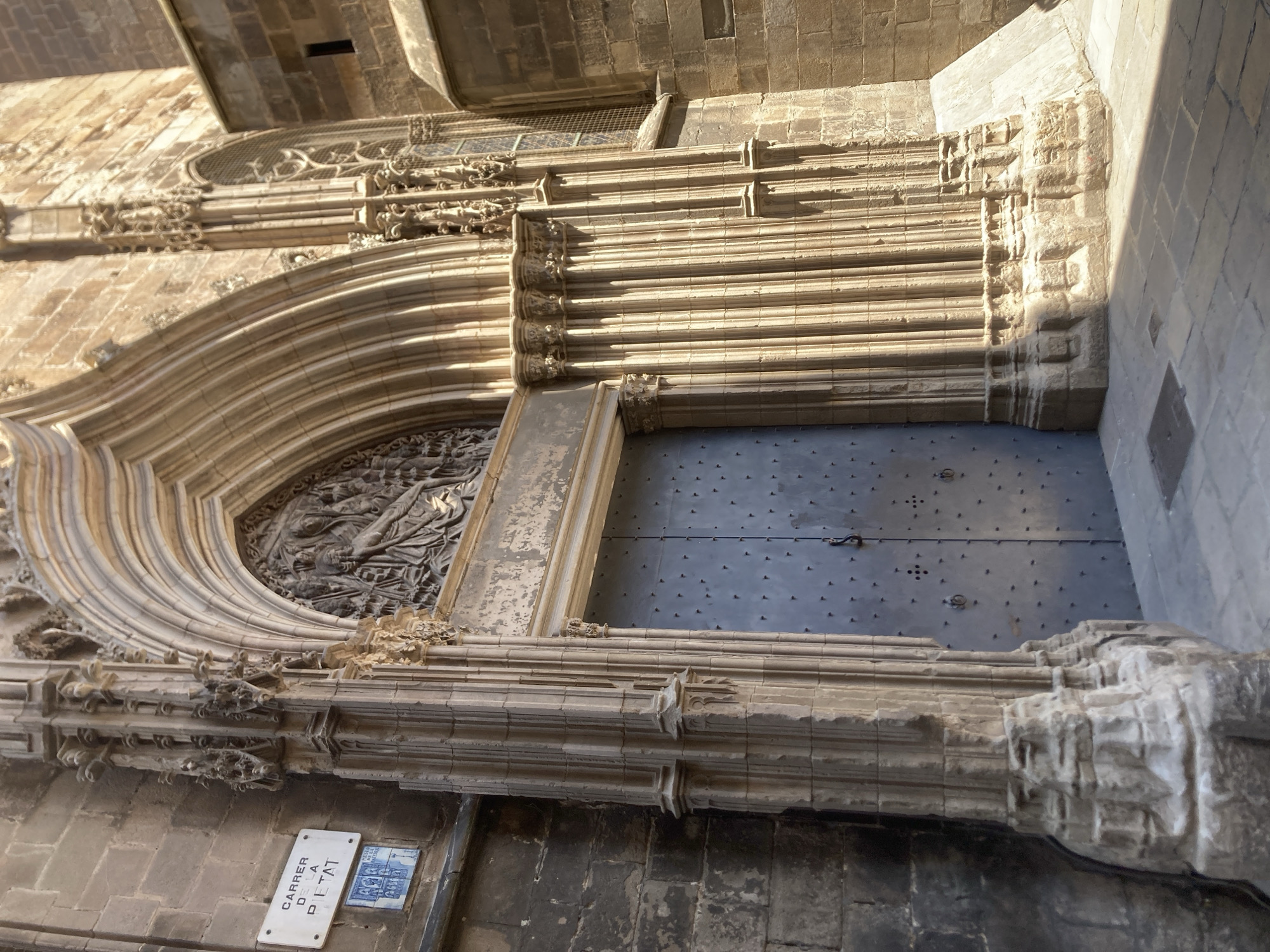 The image shows a close-up, low-angle shot of a grand arched stone doorway of a historic building in Barcelona, Spain. The doorway is flanked by intricately carved stone columns with Gothic architectural elements. A large, dark metal door with numerous studs and two ring handles is visible in the center of the frame. Above the door, a detailed bas-relief sculpture depicts a religious scene. To the left of the doorway, a white street sign with blue lettering reads "CARRER DE LA PIETAT" and a small blue tiled plaque with religious figures is mounted on the stone wall. The ground in front of the doorway is paved with stone tiles, and sunlight casts strong shadows. The overall impression is one of grandeur and historical significance.