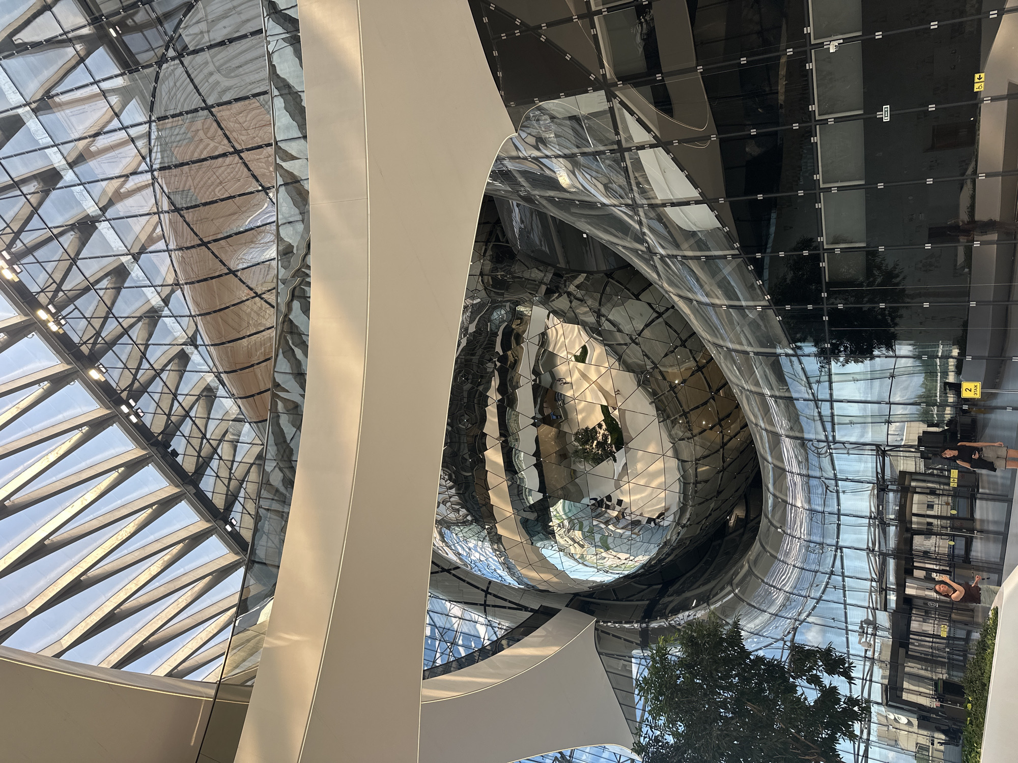 The image shows the interior of the new terminal of Pulkovo Airport in Saint Petersburg, Russia. The terminal features a unique architectural design with a large, spherical glass structure at its center. Sunlight streams in through the glass ceiling and walls, reflecting off the metallic surfaces and creating a bright, airy atmosphere. In the foreground, two women are standing near the entrance, and a tree adds a touch of greenery to the scene. The overall impression is one of modern, innovative design and a welcoming, spacious environment.