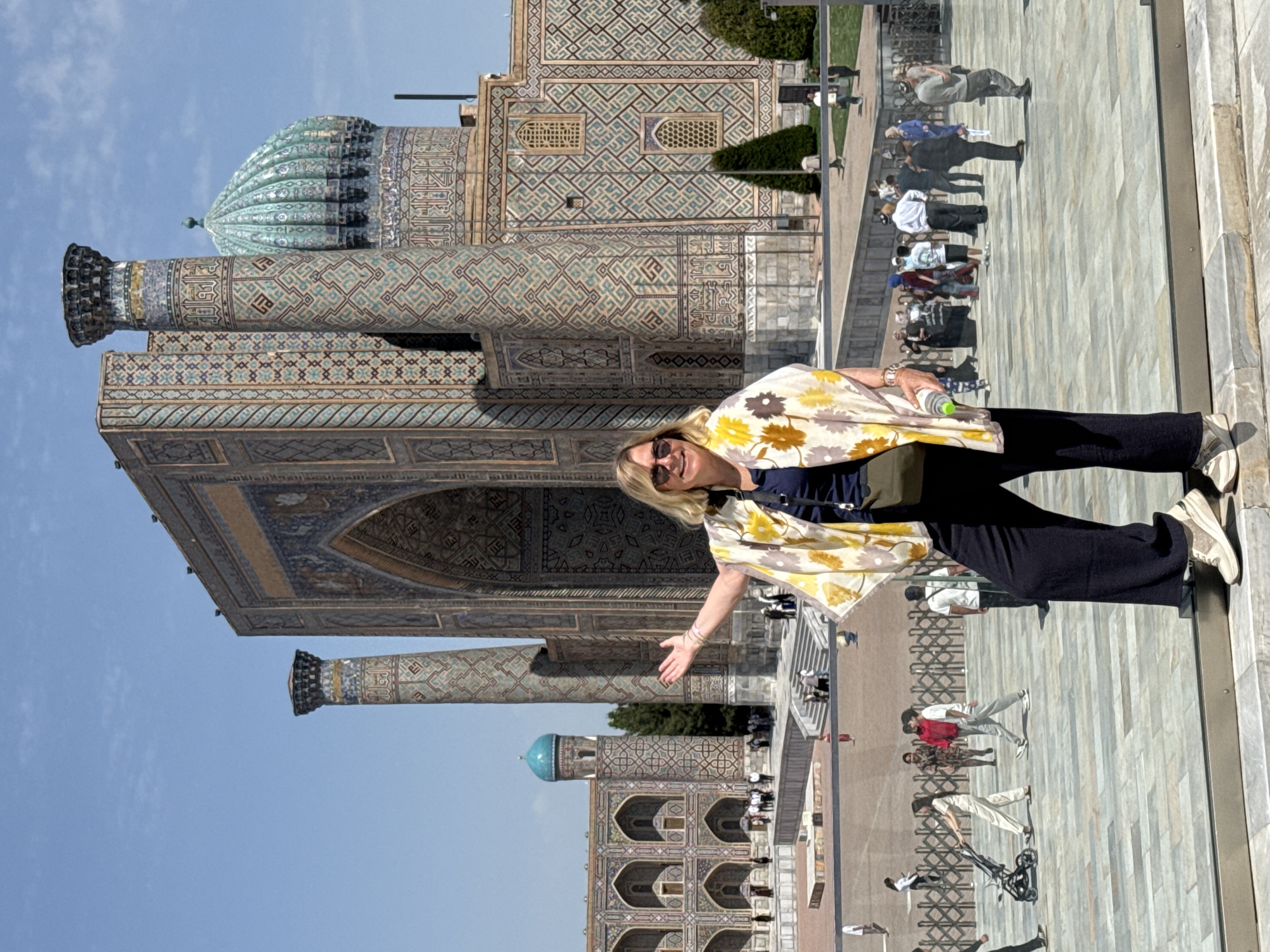 The image shows a woman posing in front of the Registan Square in Samarkand, Uzbekistan. The woman, with blonde hair and sunglasses, is wearing a floral shawl and dark clothing. She is positioned in front of a reflective surface, presumably glass or polished stone, which mirrors the surrounding architecture and other people. The architecture includes a large building with a turquoise dome and intricate mosaic tilework, with smaller structures also displaying similar tilework. Other people are visible in the background and reflected in the surface, suggesting a public space bustling with activity. The sky is clear and blue, indicating a sunny day.
