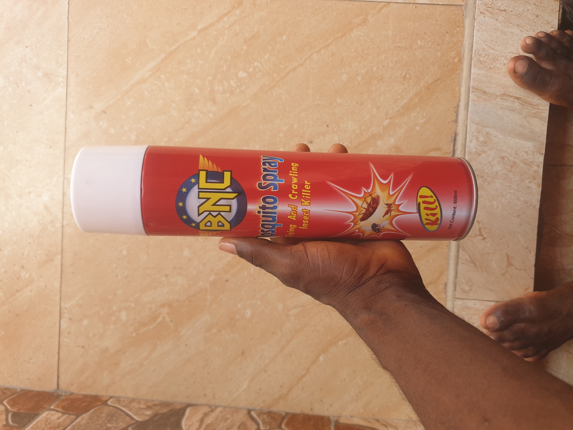 In Jalingo, Nigeria, an indoor scene depicts a dark-skinned individual's right arm and hand holding a red BNC brand aerosol can. The can, labeled "Mosquito Spray Flying And Crawling Insect Killer," features a white cap and images of a cockroach, fly, and mosquito within an impact burst, along with the text "KILL!" and "Net Content: 600ml." The individual's left foot and partial right foot are visible on the light brown tiled floor. A dark grout line traverses the floor horizontally. To the left, a textured wall section composed of irregular, multi-toned brown and orange tiles adjoins the lighter tiled surface.