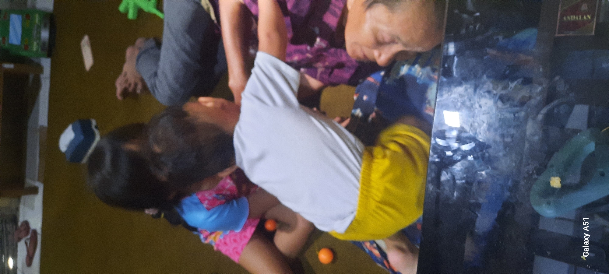 The image depicts a family scene indoors, likely in a residential setting within Ploso, Indonesia. In the foreground, a young child wearing yellow shorts and a white t-shirt is positioned centrally, facing away from the camera and towards another individual. To the left, another child, possibly a sibling, is partially visible, wearing a pink and blue patterned top. An elderly person, appearing to be a grandparent, is lying down or seated on the floor, with their head turned towards the children. The floor is a brown, carpet-like material. Scattered on the floor are small orange spherical objects, possibly toys. In the background, there are glimpses of household furniture, including what appears to be a wooden cabinet or table. Further in the background, a green toy vehicle and a blue and white object, possibly a tissue box, are visible. The overall impression is one of a casual, domestic interaction, with multiple generations present.