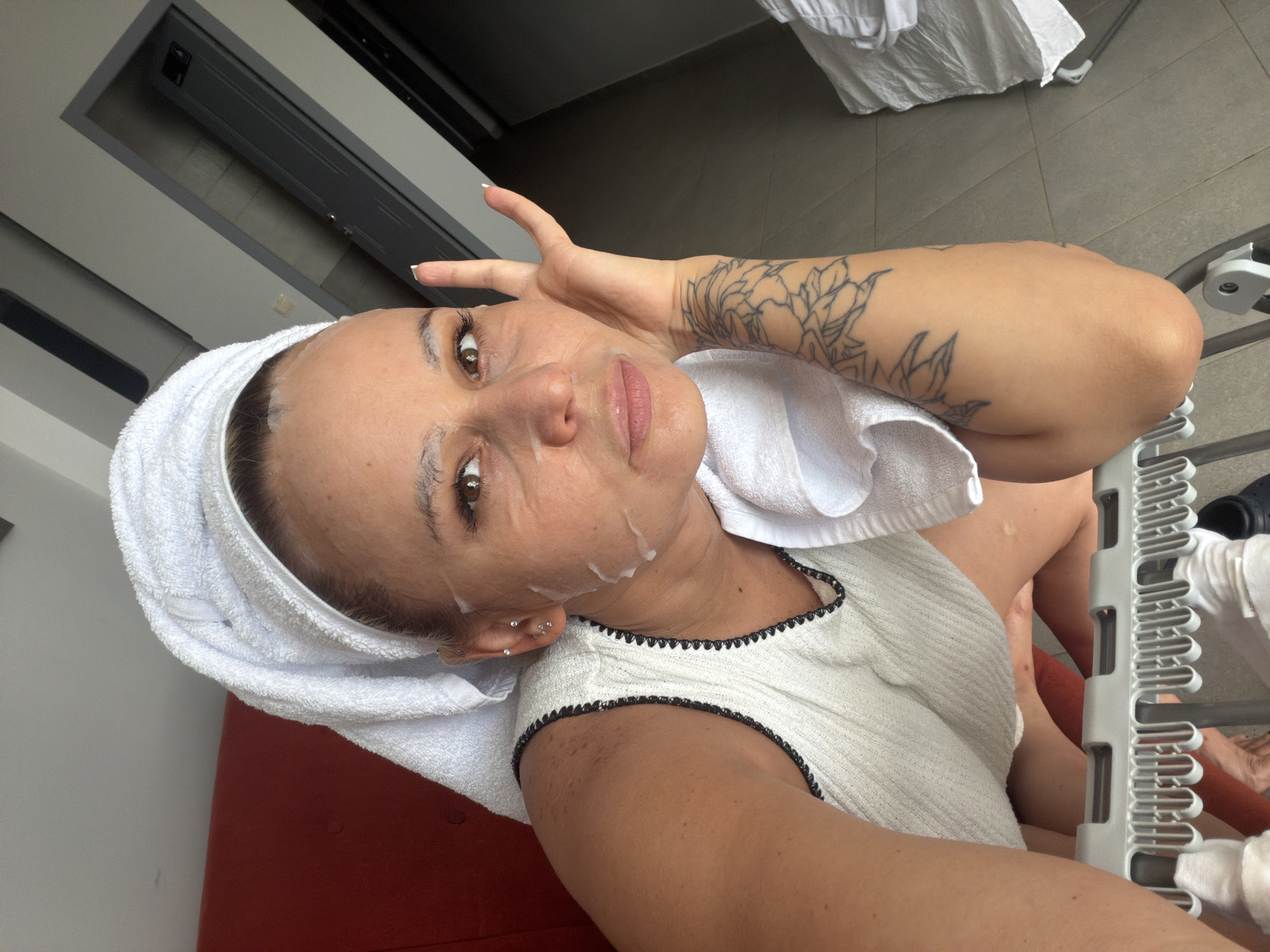A woman with a towel wrapped around her head and a facial mask on her face is shown from a high angle. She is wearing a white sleeveless top with black trim and has a tattoo on her arm. She is touching her face with her hand. The background includes a red couch, a grey door, and tiled flooring. The scene appears to be indoors. The provided crops hint at a bathroom or bedroom setting, but the specific location in Sarapul, Russia, is not discernible from the image itself.