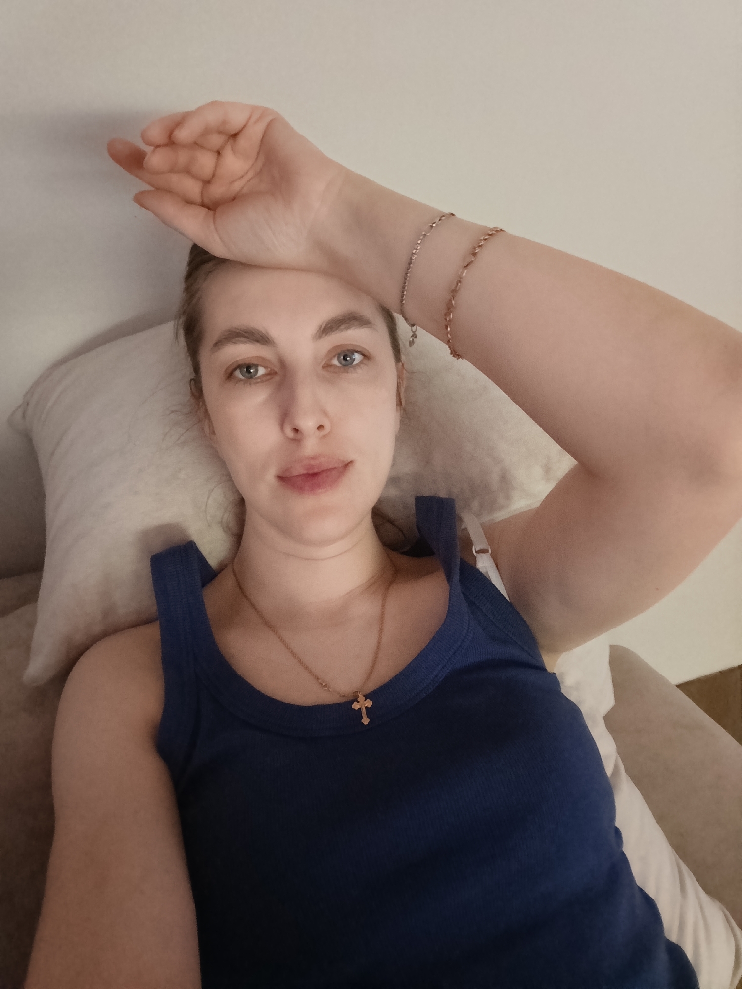 A young woman with light skin, light eyes, and light brown hair is depicted in a close-up, high-angle shot, lying down indoors. She is positioned on her back, resting her head on a light-colored pillow, which is placed on a bed or sofa. Her gaze is directed towards the camera with a neutral to slightly pensive expression.
She is wearing a dark blue ribbed tank top. A gold-colored chain necklace with a small cross pendant is visible around her neck. On her left wrist, she wears two delicate gold-colored chain bracelets. Her left arm is bent, and her hand rests gently on her forehead, obscuring a portion of her hairline and forehead. The background behind her is a plain, light-colored wall. The lighting in the image is soft and even, suggesting an indoor setting during the daytime or in a well-lit room.
The image conveys a sense of rest or relaxation. No specific objects or details in the immediate surroundings overtly indicate the location as Chaikovsky, Russia, beyond the context provided. There is no visible text in the image.