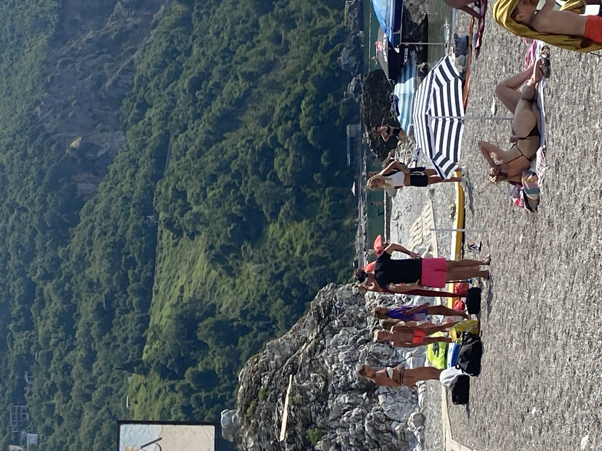 In Prijevor, Montenegro, on a sunny day, a pebble beach is crowded with people enjoying the coast. The beach is situated at the base of a steep, lush green mountain that dominates the background, with scattered buildings visible on its slopes. The water is calm and clear, with several boats docked in the distance.
In the foreground, a woman in a black bikini reclines on a towel under a striped umbrella, shielding her face from the sun. To her right, a child wearing an orange towel draped over their shoulders stands partially visible. Closer to the left, a group of people, including adults and children, are gathered near a rocky outcrop. Some are wearing swimwear, and beach bags and yellow floatation devices are scattered around them. Further back, several beach umbrellas, some with printed logos, are set up. The scene captures a typical summer day at a popular coastal destination, with a blend of relaxation and activity.