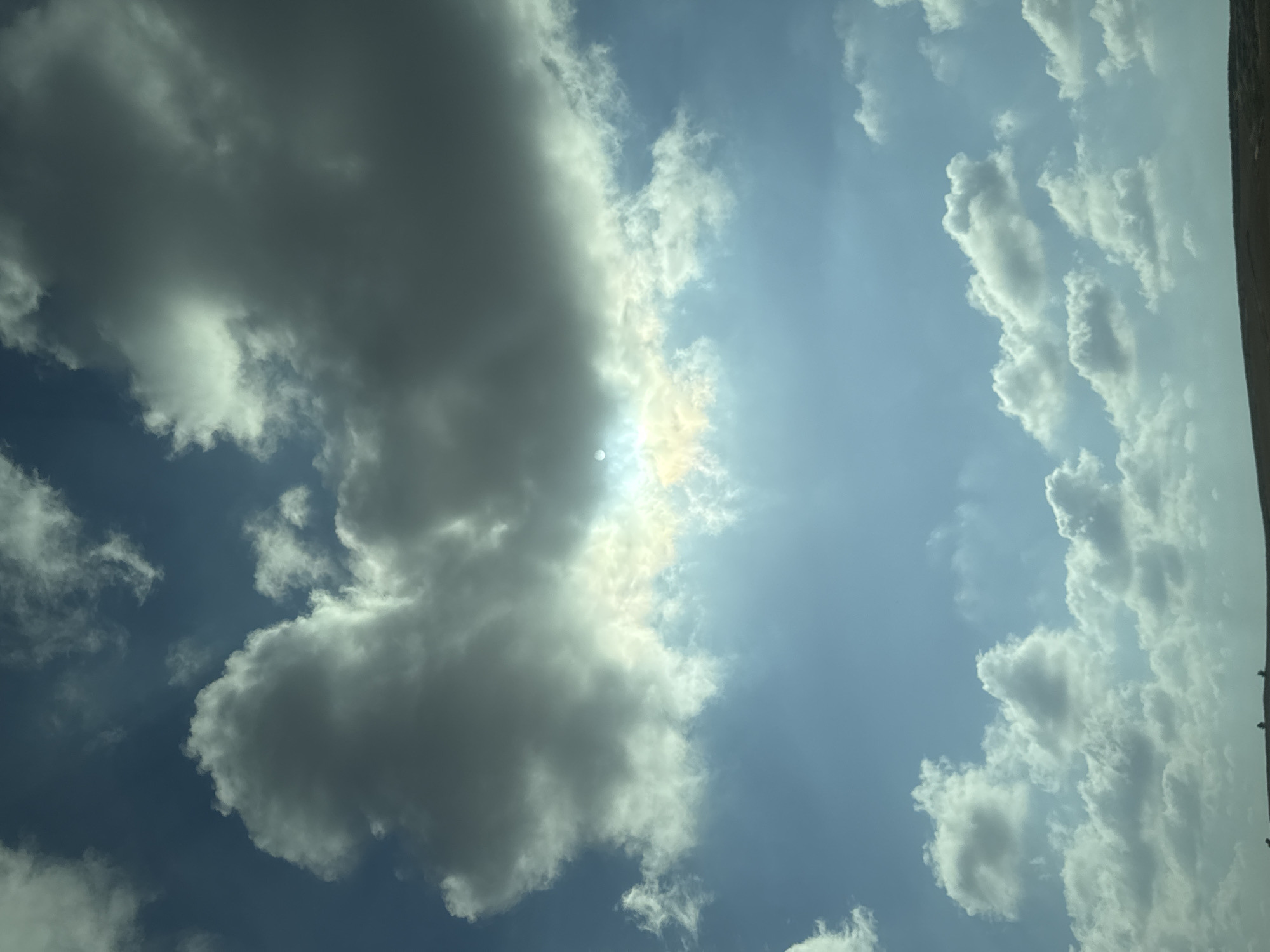 The image shows a bright, daytime sky with a mix of cumulus clouds. A large, dense cloud formation is on the left side of the frame, partially obscuring the sun. The sun's rays are visible through the lighter parts of the cloud. The right side of the image displays a lighter blue sky with smaller, scattered clouds. The top-right corner shows some terrestrial element, appearing like a tree line or distant hill. The location is Yoav Regional Council, Israel.