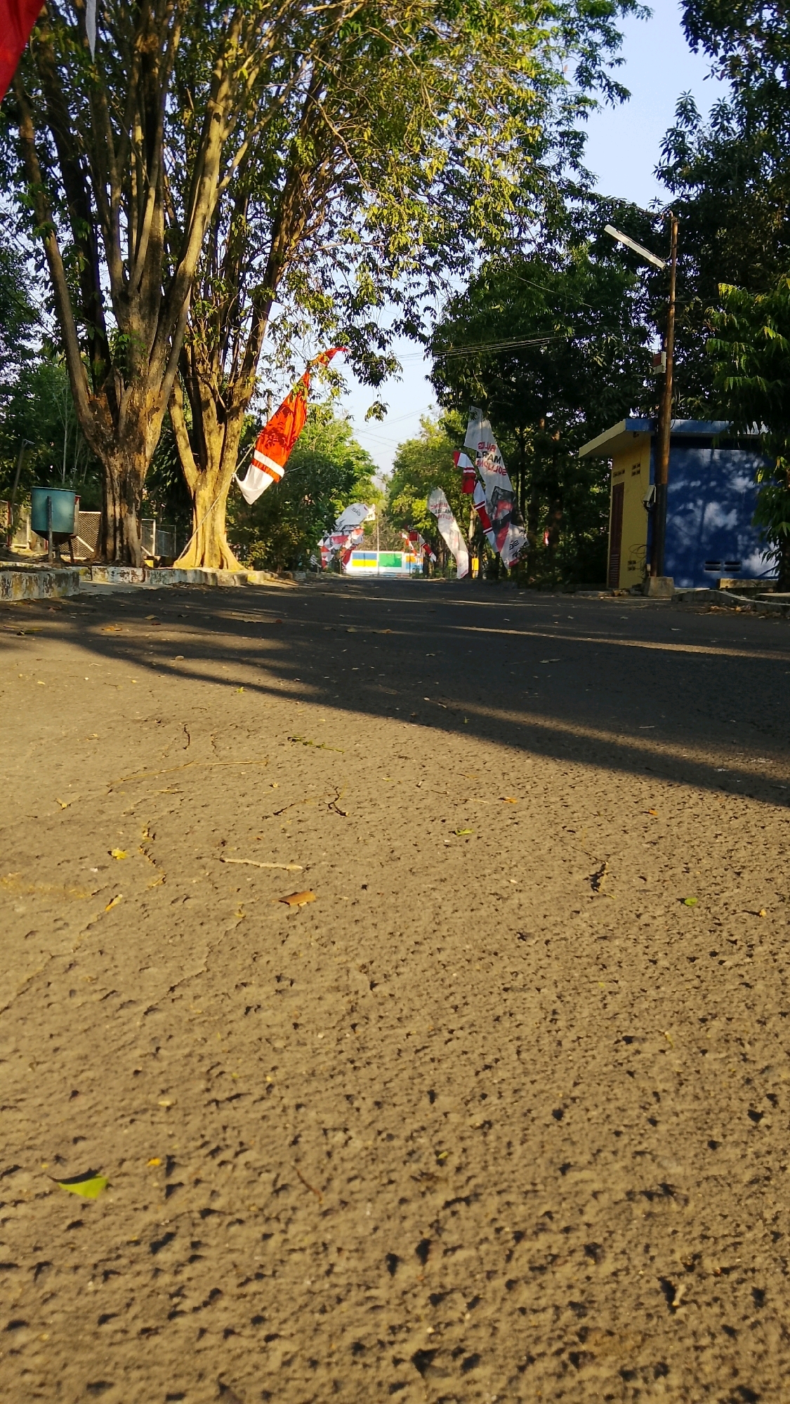 The image depicts a low-angle shot of an asphalt road in Tlogobendung, Indonesia, on a bright, sunny day. Trees with dense green foliage line the sides of the road, casting long shadows across the asphalt. Several red and white flags, some with printed text, are suspended from poles or trees along the road. In the background, a small yellow building with a blue section and a blue fence are visible, partially obscured by trees. The road surface is rough and shows signs of wear, with small stones and debris scattered across it. A light pole with a solar panel is positioned near the yellow building. The sky above is clear and blue.