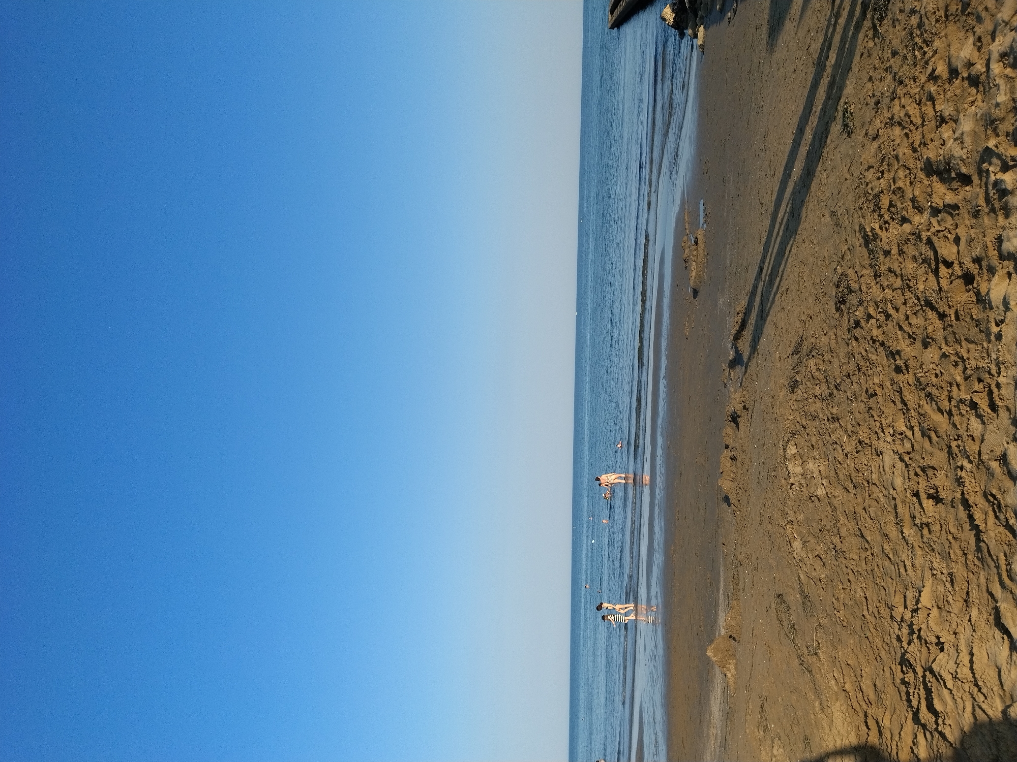 This image captures a serene beach scene in Caorle, Italy, under a clear blue sky. The foreground is dominated by wet sand, showing ripples from receding waves and the long shadows of unseen individuals. Further back, shallow water laps at the shore, reflecting the sky. In the mid-ground, several people are visible wading or standing in the water. Two individuals, a child in a striped shirt and an adult in swimwear, are walking near the water's edge. Further to the right, a group of people are bathing, with one person standing in the water and another partially submerged. In the far distance, the calm sea stretches to the horizon, and a concrete jetty is partially visible on the right side of the frame. The overall atmosphere is one of peaceful relaxation on a sunny day.