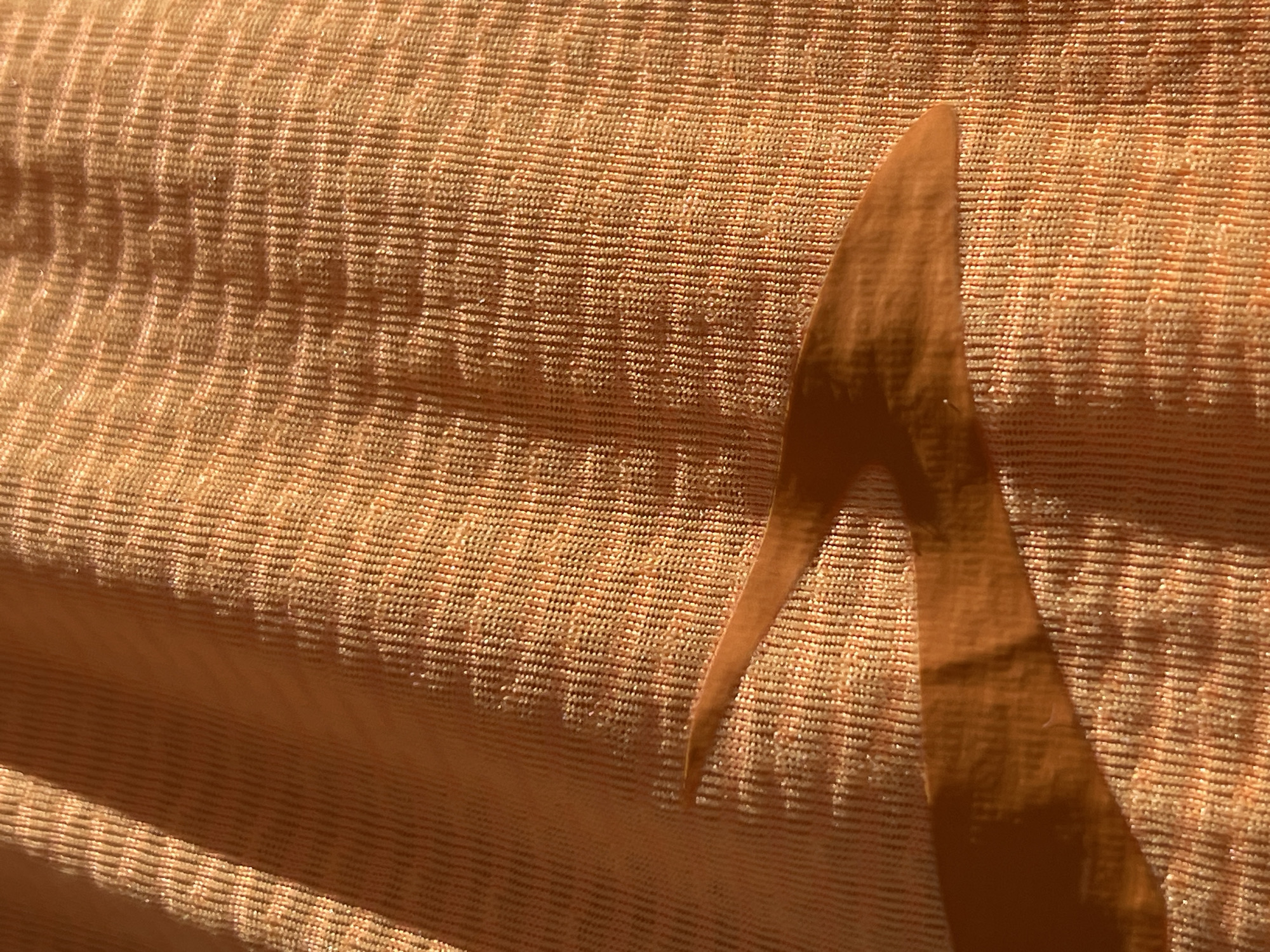 The image shows a close-up view of textured fabric with a pattern of horizontal and vertical lines, creating small square-like units. The fabric is primarily a warm, golden-brown color with variations in tone due to light and shadow. A dark, oddly shaped paper cut out with angular points and edges is situated on the fabric.