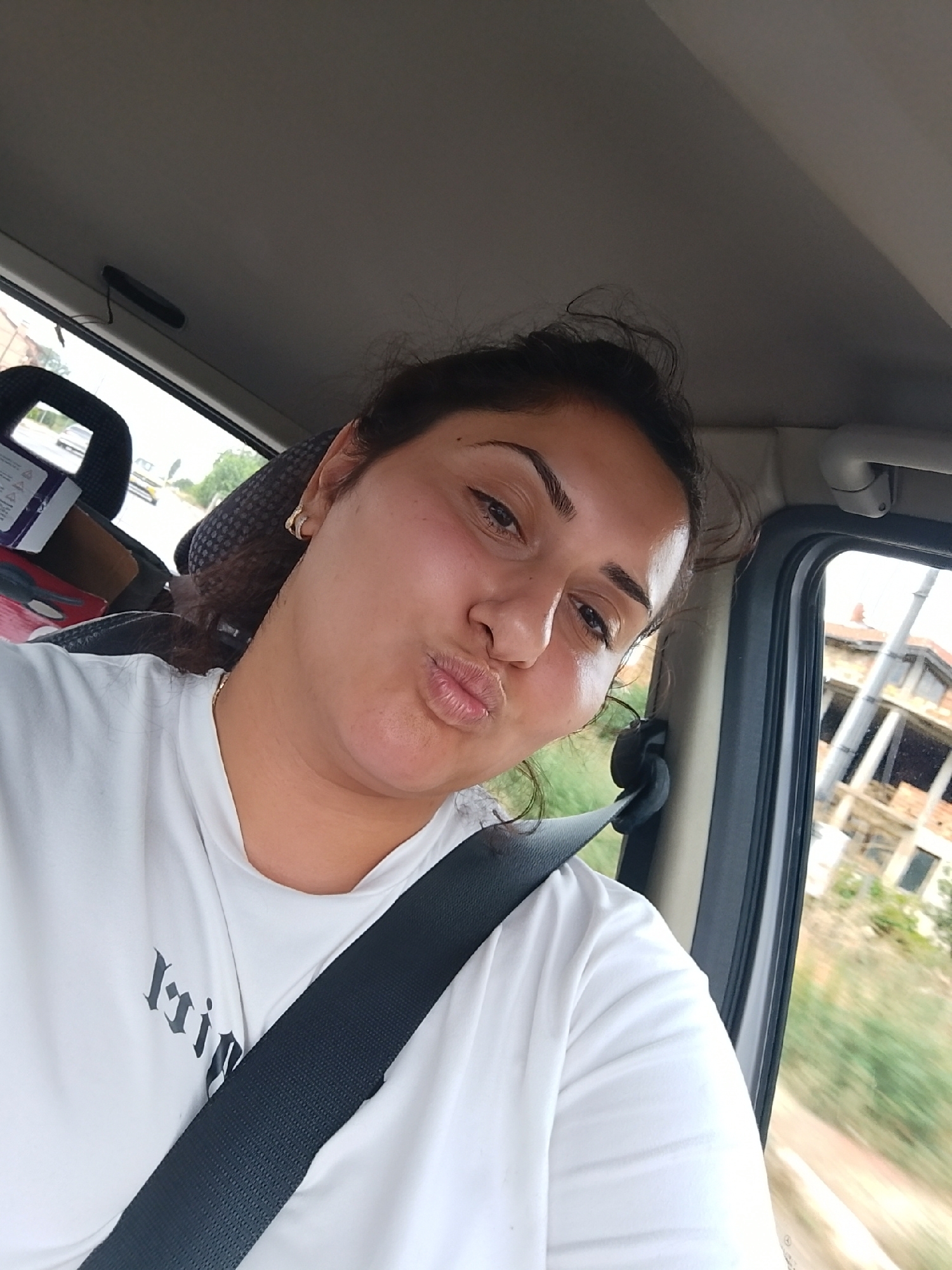 A woman with dark hair and makeup is taking a selfie in the driver's seat of a car. She is wearing a white t-shirt and a black seatbelt. The car appears to be moving, as indicated by the motion blur outside the window, which shows buildings and greenery. The location is the city of Veselinovo, Bulgaria.