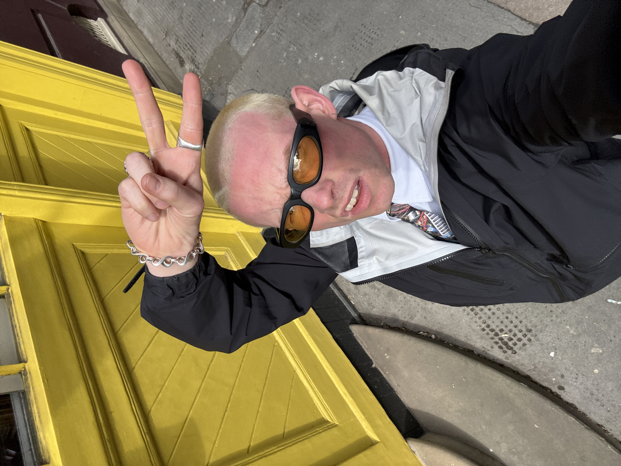A person stands on a city street in London, United Kingdom, posing for a selfie in front of a bright yellow door. The person has light skin, bleached blond hair, and wears sunglasses with orange lenses. They are dressed in a black jacket over a white shirt and are making a peace sign with their left hand, which has rings on two fingers and a silver bracelet. The background includes the yellow door, a sidewalk, and a portion of the street.