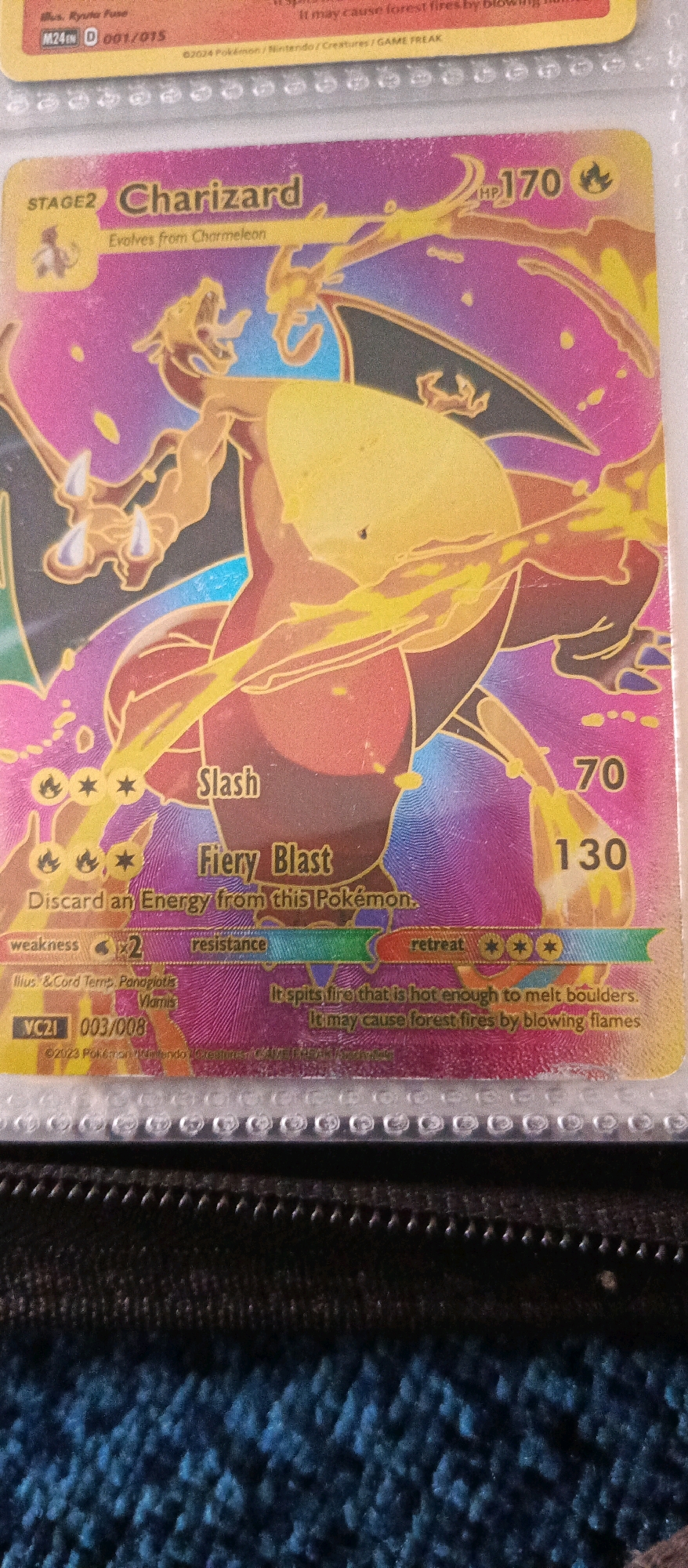 This is a close-up, eye-level photograph of a Pokémon trading card, specifically a Charizard card, displayed within a clear plastic card holder. The card itself features a holographic, shimmering finish with vibrant orange, pink, and purple hues. The central image is of the Pokémon Charizard, depicted with its mouth open and flames emanating from it. Text on the card indicates "STAGE 2 Charizard," "Evolves from Charmeleon," and its HP is "170." Below this are attack descriptions, "Slash" and "Fiery Blast," along with a cost to discard energy. Further down, it lists "weakness" and "resistance" with associated symbols and values, and "retreat" cost indicated by three energy symbols. The card also shows copyright information and a set number "VC21 003/008." The card holder is visible at the top and bottom edges, with the distinct pattern of a card binder visible behind it, suggesting the card is part of a larger collection. The background appears to be a textured, dark blue surface, possibly fabric. There are no people or any elements that place this image within the city of Brwinów, Poland.