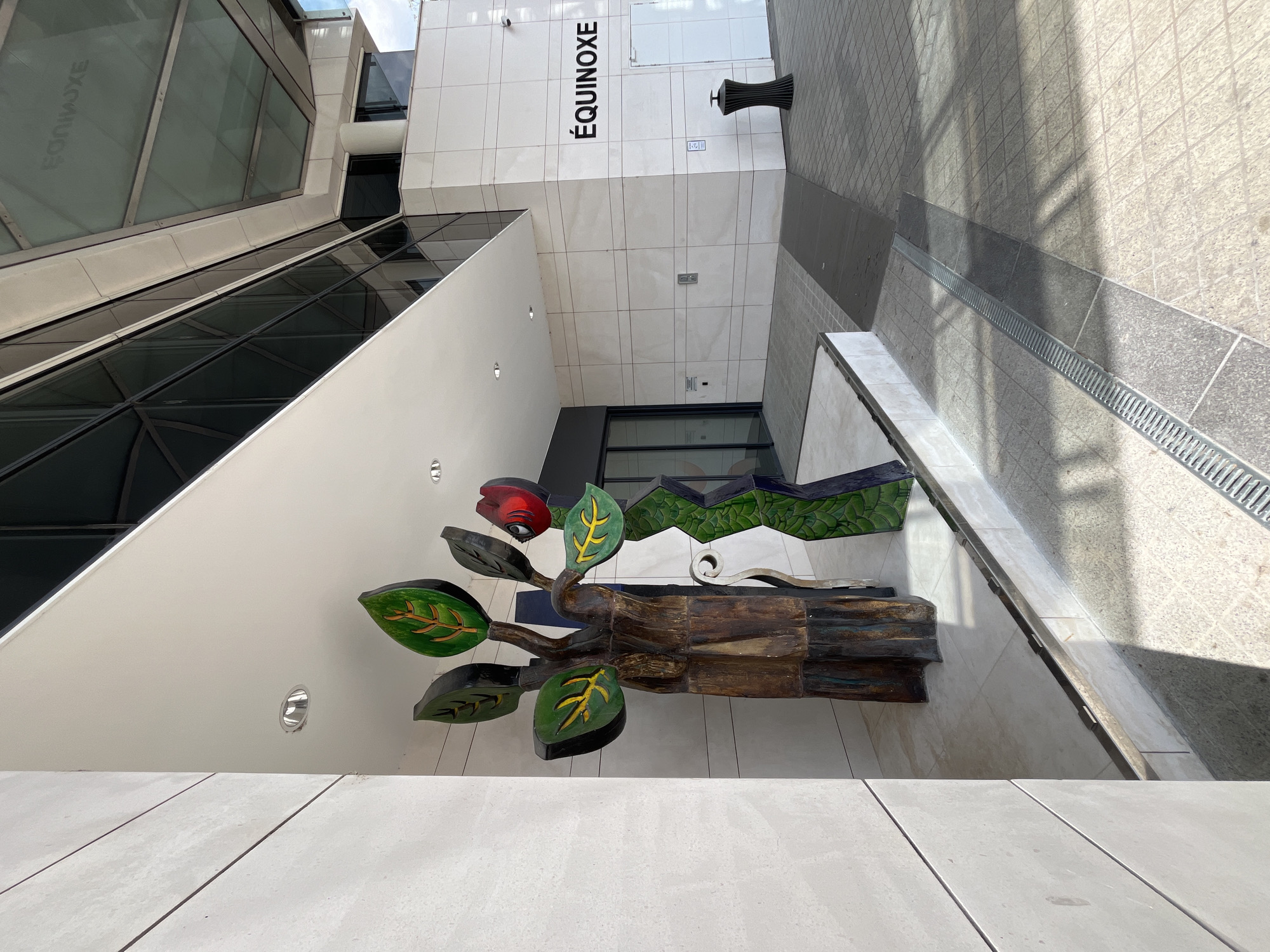 This is an outdoor, slightly high-angle, eye-level shot of a modern building entrance. The building has a white tiled facade with a large "ÉQUINOXE" sign prominently displayed. To the left, there's a contemporary glass structure with angled windows. In the foreground, a wooden sculpture of a stylized tree with colorful green leaves and a red, eye-like object is visible. A dark, ribbed waste bin stands near the "ÉQUINOXE" sign. The ground is paved with light-colored tiles and a metal grate runs along a drainage channel. Sunlight casts shadows on the tiled ground, suggesting it is daytime. The scene appears to be a public outdoor space, possibly a plaza or entrance area. There is no activity or people visible in the frame.