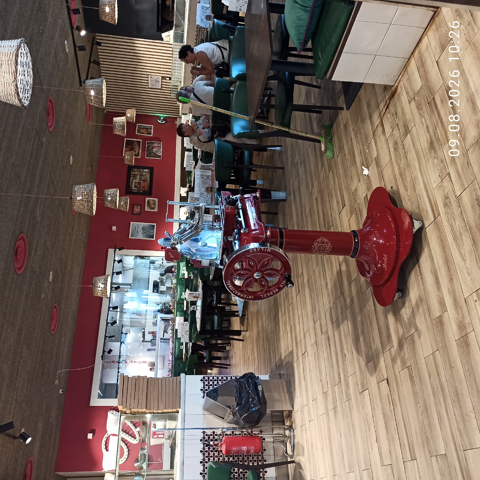 The image depicts the interior of a restaurant, specifically identifying the location as Molfetta, Italy. The scene is set with a bright red wall as a prominent backdrop, contrasting with the light wood-paneled floor. A vintage red meat slicer, labeled "INTERNATIONAL VAN BERKEL MILANO," stands as a central object in the foreground.
Several tables with green chairs are arranged throughout the restaurant, with some occupied by patrons. In the mid-ground, a group of three individuals are seated at a table, engaged in conversation. Two women and one man appear to be enjoying a meal or drinks. The woman on the left is wearing a striped shirt, while the man in the middle wears a white t-shirt. The woman on the right is wearing a white top.
Other notable objects in the scene include a fire extinguisher, a trash can with a black garbage bag, stacks of cardboard boxes, and several woven pendant lights hanging from the ceiling. Framed pictures adorn the red wall, adding to the restaurant's decor. A broom with green bristles leans against a table, suggesting recent cleaning or preparation. The overall ambiance is casual and lively, indicative of a dining establishment.
The image is dated August 9, 2026, at 10:26 AM.