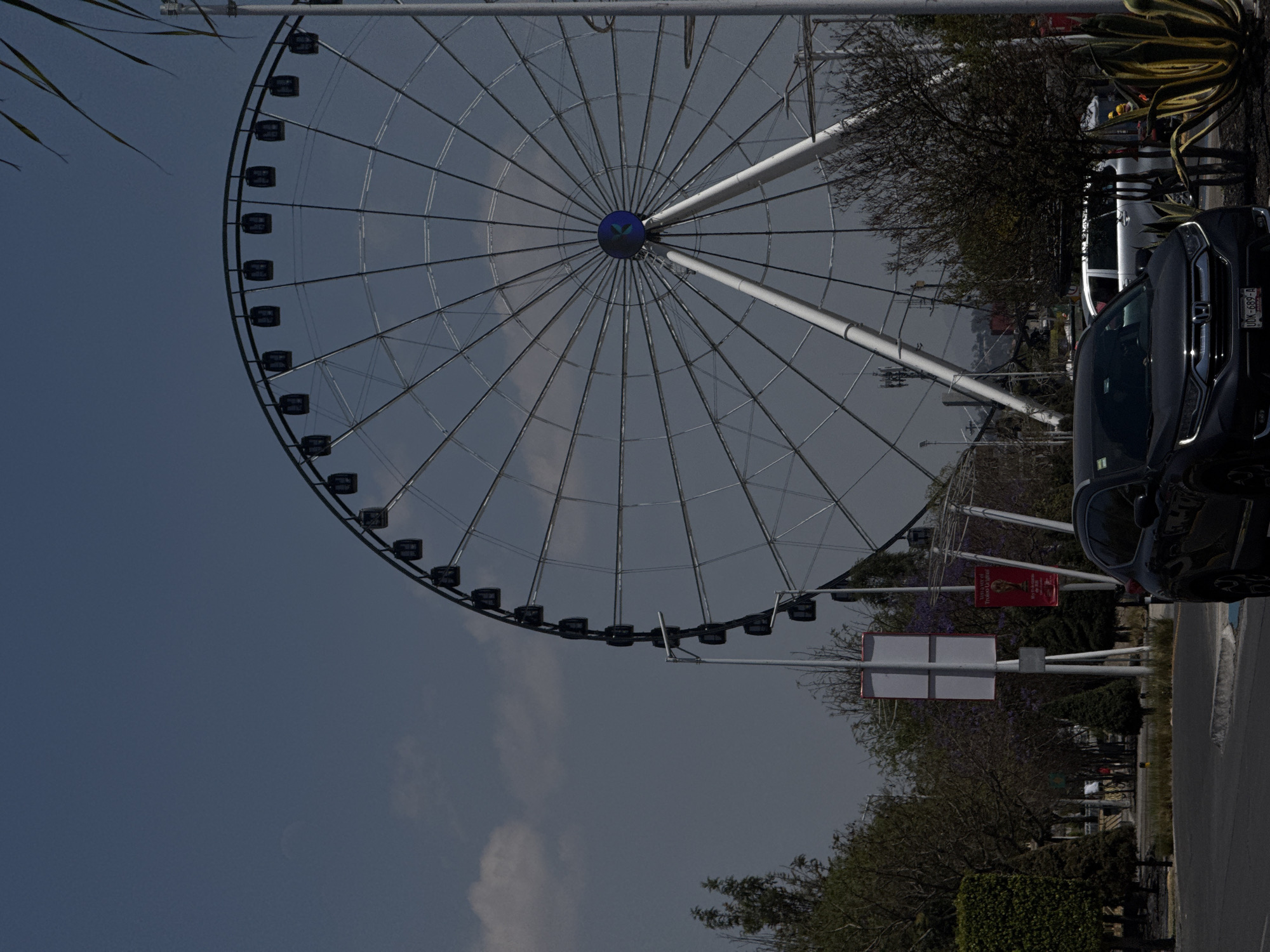 The image displays an outdoor daytime scene in Puebla, Mexico, centered on the Estrella de Puebla Ferris wheel located in the Angelópolis district, near the Parque Lineal. The Ferris wheel dominates the right side of the frame, featuring a white support structure, a dark-grey wheel frame with numerous spokes, and approximately 30 dark gondolas. The central hub is blue with a lighter blue stylized "K" or starburst design. The sky is light blue with scattered white clouds; a faint crescent object, likely the moon, is visible in the upper left quadrant.
In the immediate foreground, a dark-colored Honda CR-V SUV is parked or stopped on an asphalt road. Its front license plate, "UDK-689-A," is visible. A silver SUV is partially visible behind the Honda to the right. The road features a light grey curb. Various trees and bushes line the left side of the road and extend into the mid-ground, some exhibiting purple blossoms. In the lower right, several large, variegated agave plants with yellow and green leaves are visible, alongside smaller, bare-branched trees. Multiple grey street light poles and utility poles are distributed throughout the scene. A tall white signpost with two blank rectangular panels stands in the mid-ground, behind which a red banner with text "Vas a ver el Titulo Original" and a trophy graphic is partially visible. No individuals are depicted. The scene is illuminated by natural sunlight.