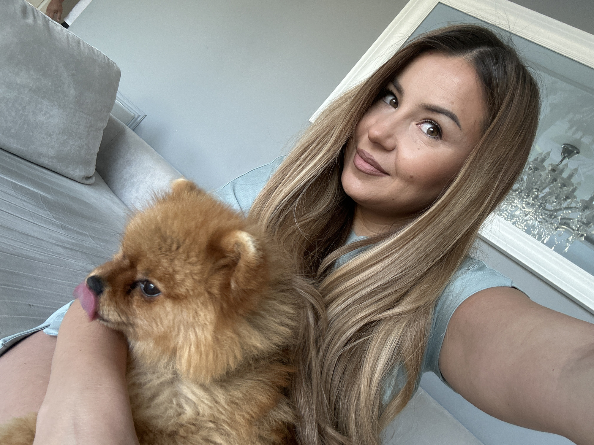 A woman is taking a selfie while holding a small, fluffy, ginger-colored dog on her lap. The dog is licking the woman's arm. The woman has long, blonde-brown hair and is wearing a light blue shirt. She is sitting on a light grey couch. In the background, there is a large, ornate mirror with a crystal chandelier visible in its reflection. The overall environment appears to be a well-lit indoor space, possibly a living room. The location is in Asenovgrad, Bulgaria.