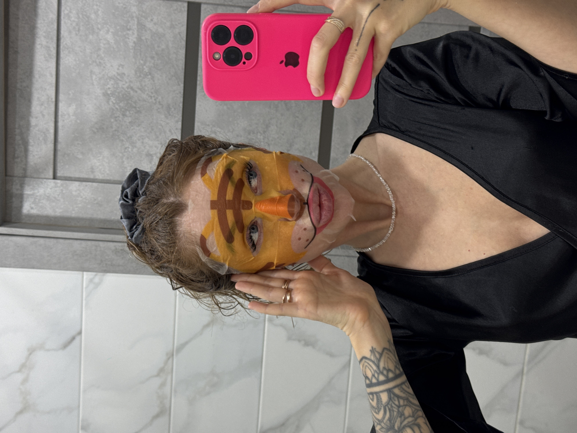 A person is taking a selfie in front of a bathroom tiled with white marble-patterned tiles, and gray-textured tiles with a vertical seam. The person is wearing a black top, a silver necklace, and several rings. The person is holding a pink iPhone in their right hand, and their left hand is near their face. The person has a face mask on with a tiger design, including an orange nose. The person has tattoos on their right forearm and left hand. The location is inside a bathroom; there is no information to indicate that this is in the city of Chaikovsky, Russia.