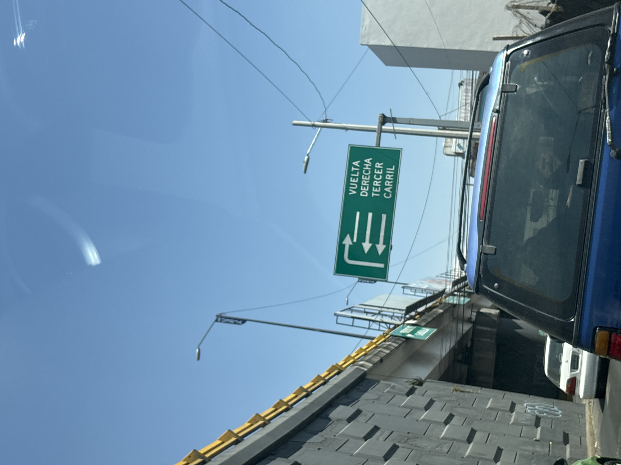 The scene depicts an urban road environment under a clear blue sky, viewed from within a vehicle, likely in traffic, at the **Distribuidor Vial 5 de Mayo in Puebla, Mexico**.
In the foreground, the rear section of a dark blue SUV is visible, including its rear window, roof rack, and a portion of its red taillight bar. Ahead and slightly to the left of the SUV, a white sedan is partially visible. Further to the far left, a glimpse of a green vehicle is present.
To the left, a tall, grey concrete retaining wall with a patterned, block-like texture supports an elevated roadway. A yellow metal guardrail runs along the top edge of this elevated structure. A street light pole with a banner partially displaying "PUEBLA" is mounted on the elevated section. Below the elevated road, a shadowed area suggests an underpass or tunnel entrance.
In the mid-ground, a green rectangular overhead sign, mounted on a metal pole, displays white text and arrows. The text reads "VUELTA DERECHA TERCER CARRIL," indicating "RIGHT TURN THIRD LANE." Three white arrows are shown; the bottom one points sharply right, while the two above it point less sharply right.
To the right of the sign, another street light pole is visible, along with multiple utility wires crisscrossing the upper portion of the frame. A white building or structure is partially visible in the far right background, behind the utility lines. The overall environment suggests a complex urban interchange with multi-level roadways.