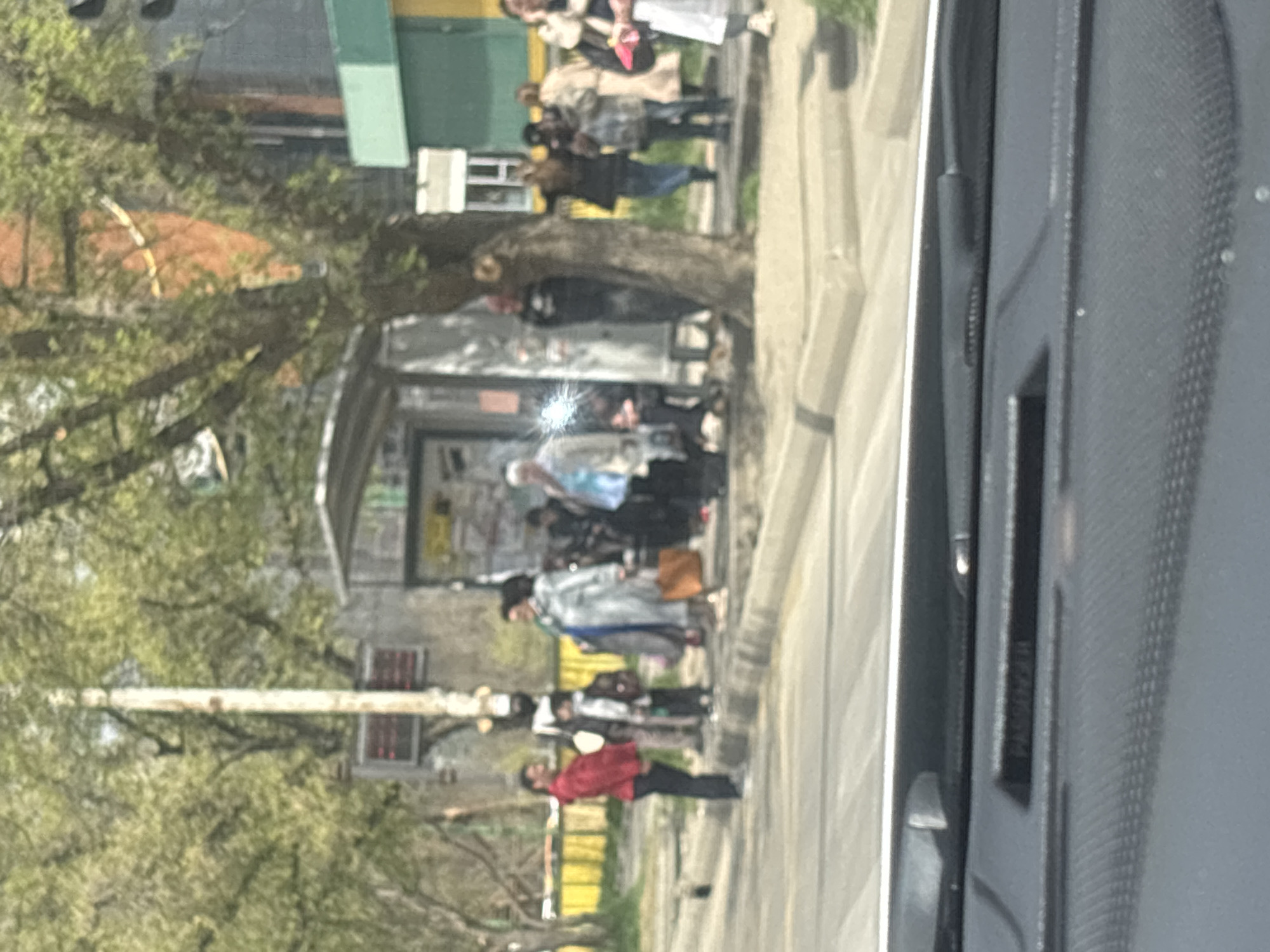 A somewhat blurred image taken from inside a car shows a group of people waiting at a bus stop in Tbilisi, Georgia. The bus stop is situated on a sidewalk lined with trees showing new spring growth. Several individuals are standing and waiting, with a mix of clothing colors and styles. One person is wearing a red shirt, while others are clad in more muted tones. The bus stop structure is basic, likely metal with some informational posters on the back. Background details include buildings of varying heights and styles, suggesting an urban setting. The inside of the car is visible on the right side of the frame.