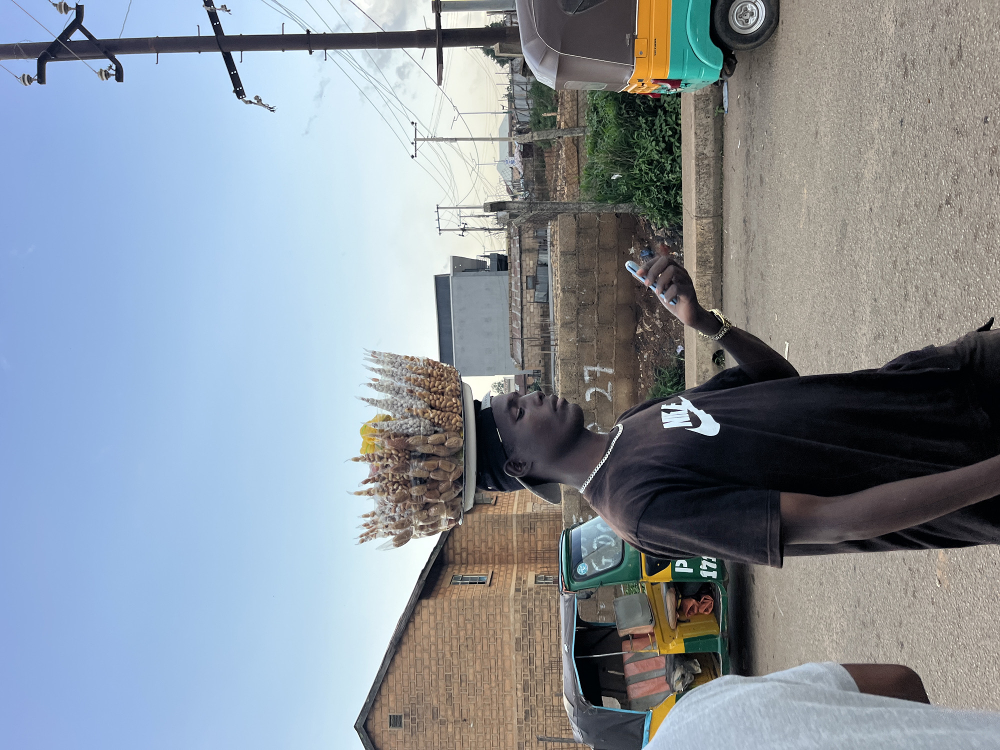 A young Black male is depicted walking on a paved street, balancing a large tray of snacks on his head. He is wearing a black t-shirt with a white Nike logo and a silver chain around his neck. He also has a gold bracelet on his left wrist. In his right hand, he holds a light blue smartphone. Behind him, a yellow and green auto-rickshaw is parked. Further in the background, a brick building and a power pole with electrical wires are visible. The sky is clear and blue. The scene appears to be in an urban environment.