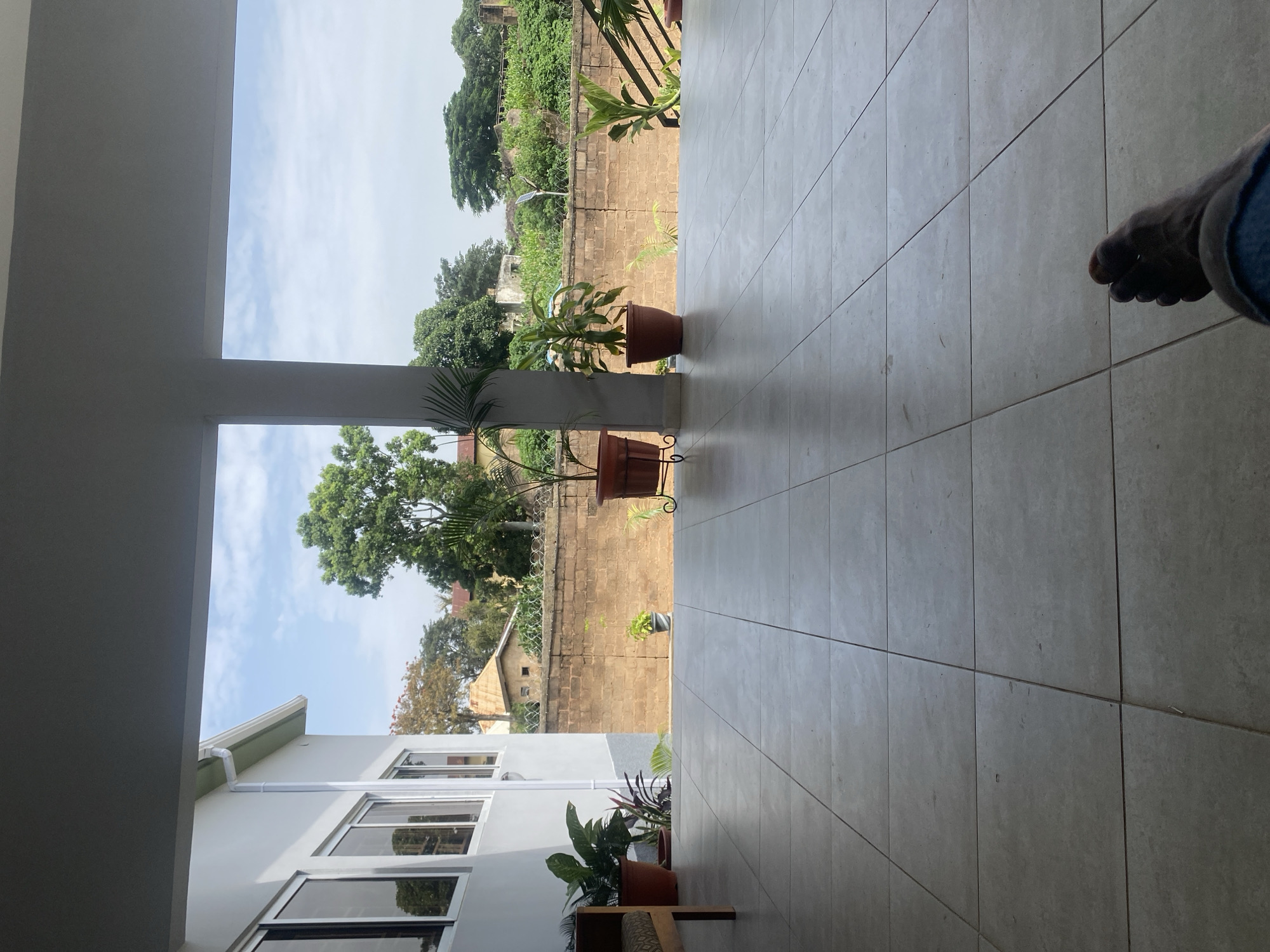 The image shows a view from a covered patio, looking out towards a backyard in the city of Jos, Nigeria. The patio is paved with light-colored square tiles. To the left, part of a white building with several large windows is visible. A wooden bench with a patterned cushion sits on the patio near the building. Several potted plants are arranged on the patio, including a large green plant in a brown pot and a smaller plant in a pot on the ground. A brick wall, topped with barbed wire, forms the boundary of the property, separating it from neighboring areas. Beyond the wall, trees and some structures are visible, suggesting a residential or semi-rural setting. The sky is partly cloudy, with blue sky and white clouds. In the foreground, a person's bare foot, with dark skin, is visible resting on the tiled floor, suggesting they are sitting on the patio. The overall impression is one of a relaxed outdoor space in a warm climate.
