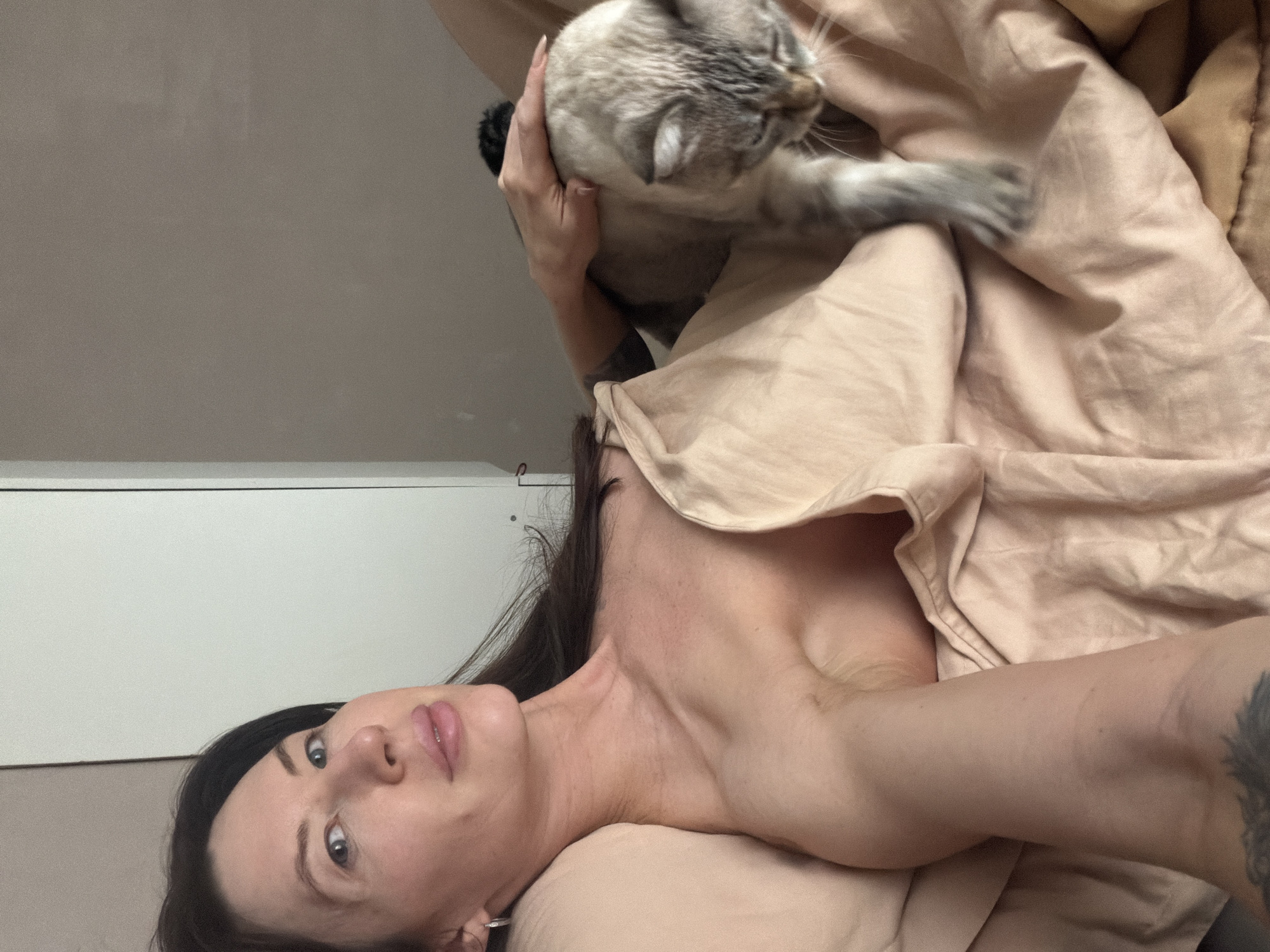 The image captures a person and a cat relaxing together in an indoor setting, likely a bedroom. The perspective is from an overhead angle, looking down at the subjects.
The main subject is a person, appearing to be a woman, lying on her back. She has dark hair, light-colored eyes, and is looking directly at the camera with a neutral expression. Her upper torso, including her shoulders and chest, is visible, partially covered by soft, light beige bedding. A tattoo is visible on her right arm.
Perched above her (when viewed upright), and held gently by her hands, is a cat. The cat has light-colored fur with darker points on its ears and face, suggesting a Siamese or similar breed. Its head is turned away from the camera, looking towards the upper right of the frame. One of its paws rests on the rumpled bedding.
The background consists of a plain, light greyish-brown wall and a white surface, possibly a headboard or shelf, along the left side. The bedding is a soft, textured light beige or tan color, adding to the cozy and relaxed atmosphere. The scene depicts a quiet, intimate moment between a pet owner and their cat. No specific time of day, weather conditions, or location cues are visible within the image itself. There is no visible text.