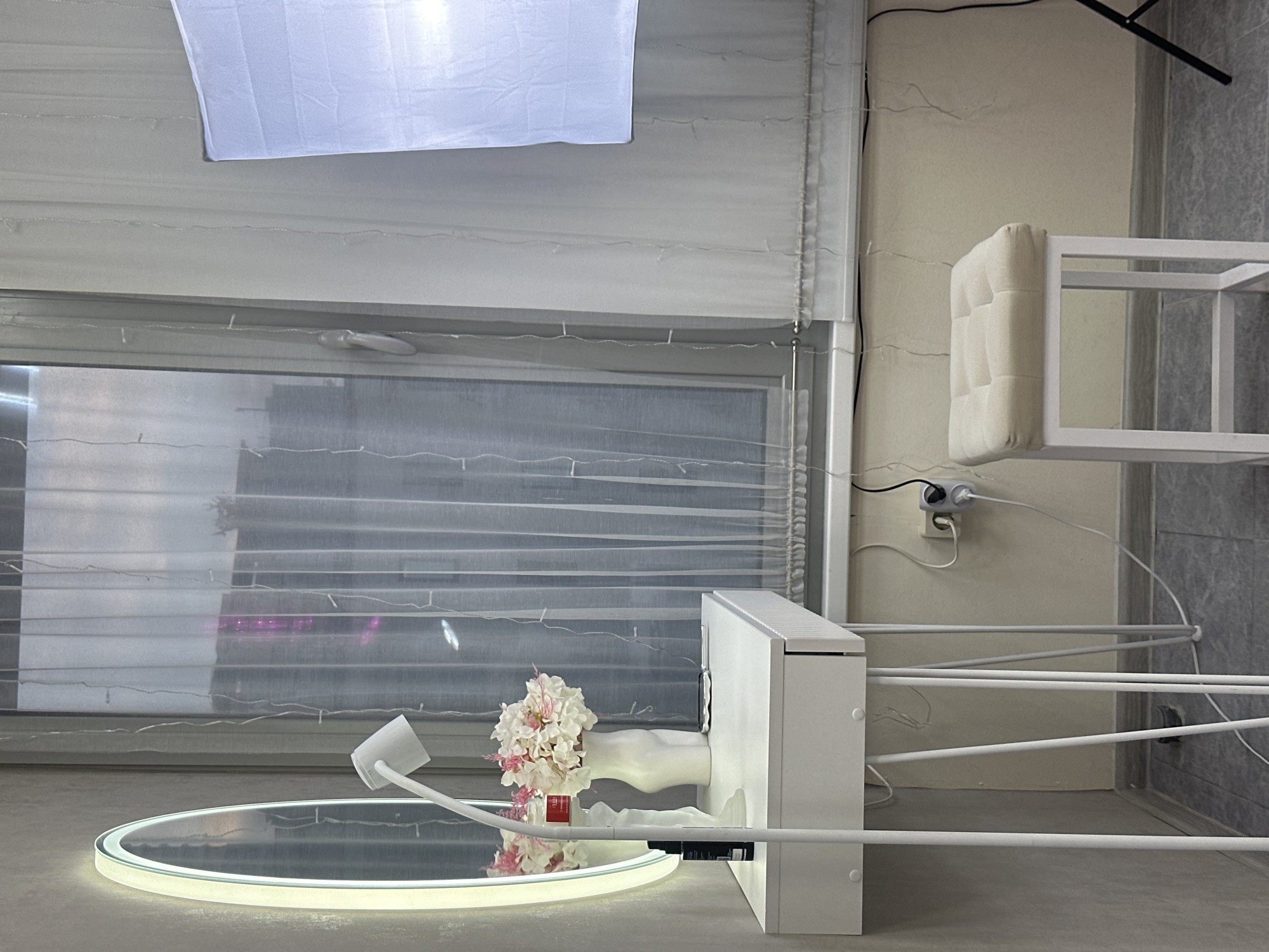 The image captures an indoor setting, possibly a studio or a room configured for product display or photography, located in Sochi, Russia. The scene is dominated by white and light gray tones, illuminated by both natural light from a window and artificial sources.
In the upper portion, a large, rectangular white softbox or light diffuser hangs, directing light downwards. Below it, a window is fitted with sheer, light-colored blinds or curtains, allowing some natural light to filter through. The window reflects elements of the room, and faint outlines of outdoor scenery are visible, along with a hint of purple light or reflection in one of the window panes.
At the center, a white, rectangular structure holds a vase filled with artificial light pink and white hydrangeas. This structure is positioned next to a round, illuminated mirror that reflects the flowers and parts of the room. A white gooseneck-style lamp or mount extends over the mirror.
To the right, a white cushioned stool or chair with a minimalist frame is visible. Further right and extending into the lower frame, a white, multi-tiered rack or shelf structure with thin bars is present, possibly a clothes drying rack. Electrical cords are visible, plugged into a wall outlet (a Schuko type, common in Russia) on a light-colored wall. A black tripod leg is partially seen in the upper right corner. The flooring is a neutral gray.
The setup suggests an activity of preparing for or conducting product photography or creating an aesthetic display. The time of day appears to be daytime, given the light from the window. A small red label with unreadable white text is visible on the vase.