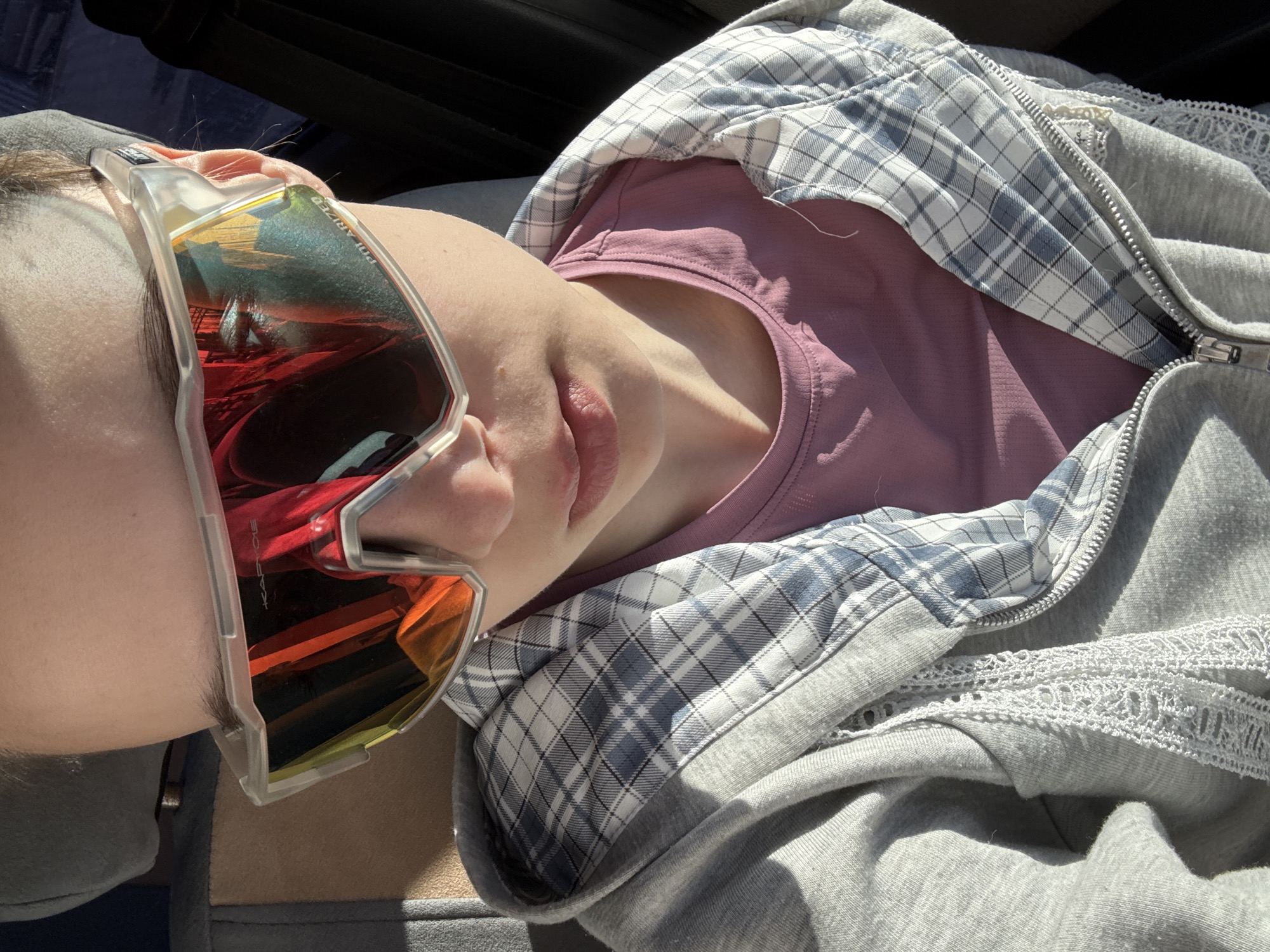 A person is lying down, wearing a grey hoodie over a mauve top and large, clear-framed sunglasses with orange and red lenses. The top layer of the hoodie has a white and blue plaid pattern. The scene seems to be inside of a vehicle. There is no verifiable evidence to place this image in Прикамский, Russia.
