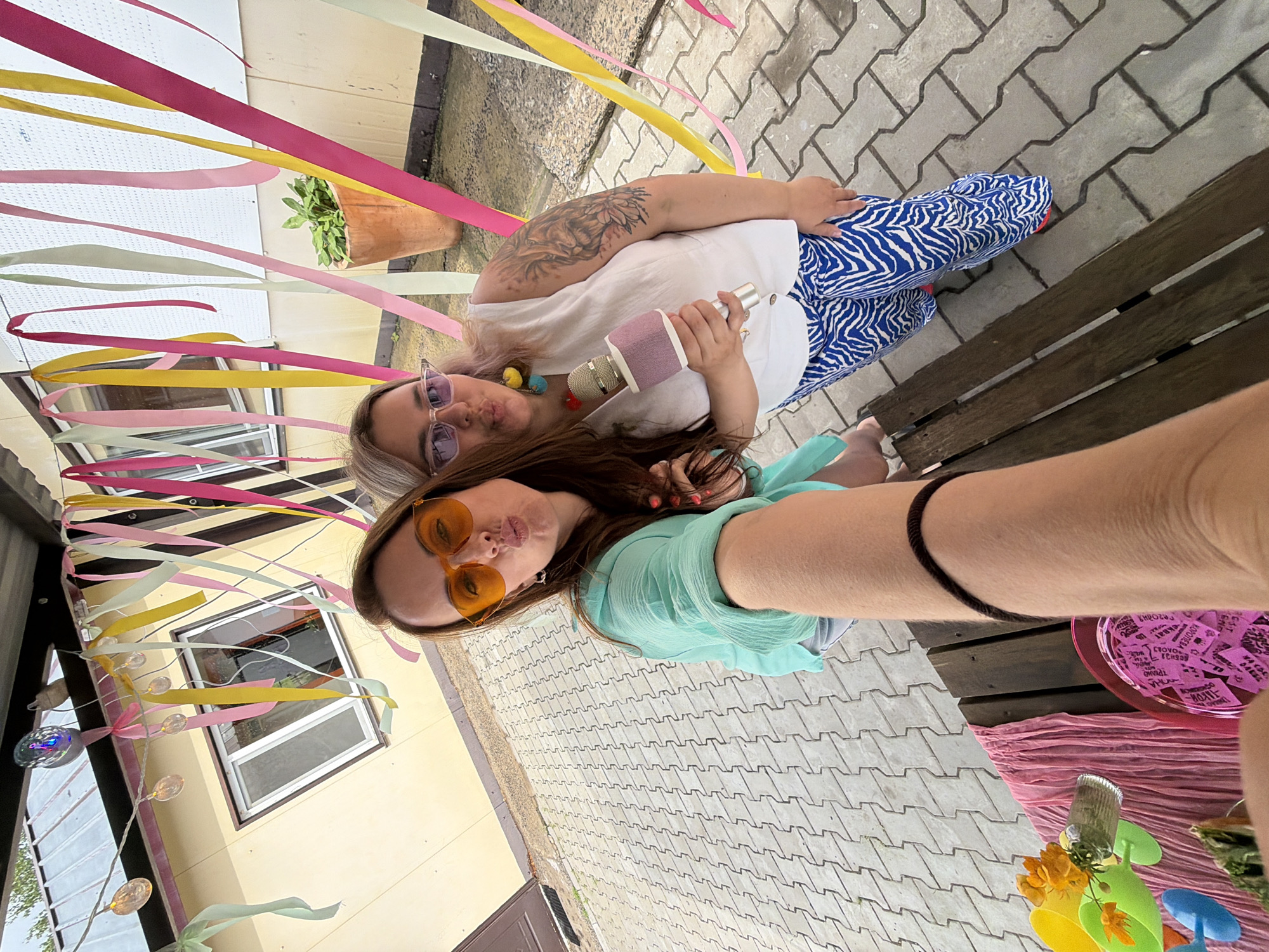 Two women are posing for a selfie outdoors. The woman on the left is wearing an aqua-colored blouse and orange sunglasses. The woman on the right is wearing a white sleeveless top and blue and white zebra-striped pants, and is holding a pink and silver microphone. They are standing in front of a yellow building decorated with colorful streamers. The ground is paved with interlocking gray bricks. There is a wooden bench to the right of the women. The scene appears to be a celebration or party. The building is situated in Birobidzhan, Russia.
