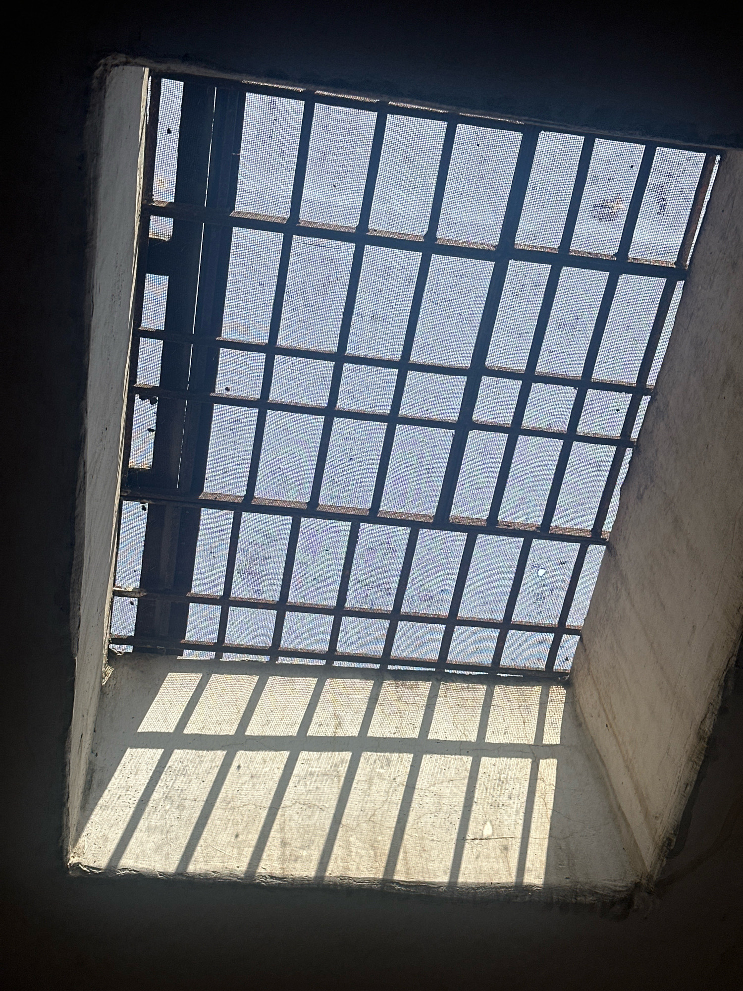 The image depicts a view looking upwards towards a window with metal bars, suggesting a confined or secure space. The window is covered with a fine mesh screen, allowing diffused light to pass through. Strong shadows cast by the bars fall across a textured surface below, likely a wall or floor, indicating a bright light source outside, possibly the sun. The surrounding structures appear to be made of concrete or plaster, with a rough texture. The overall impression is one of a utilitarian or possibly aged architectural setting. The location in Khazan Singhwala, India, implies a specific geographic and cultural context, though the image itself does not contain overt visual cues to confirm this location.