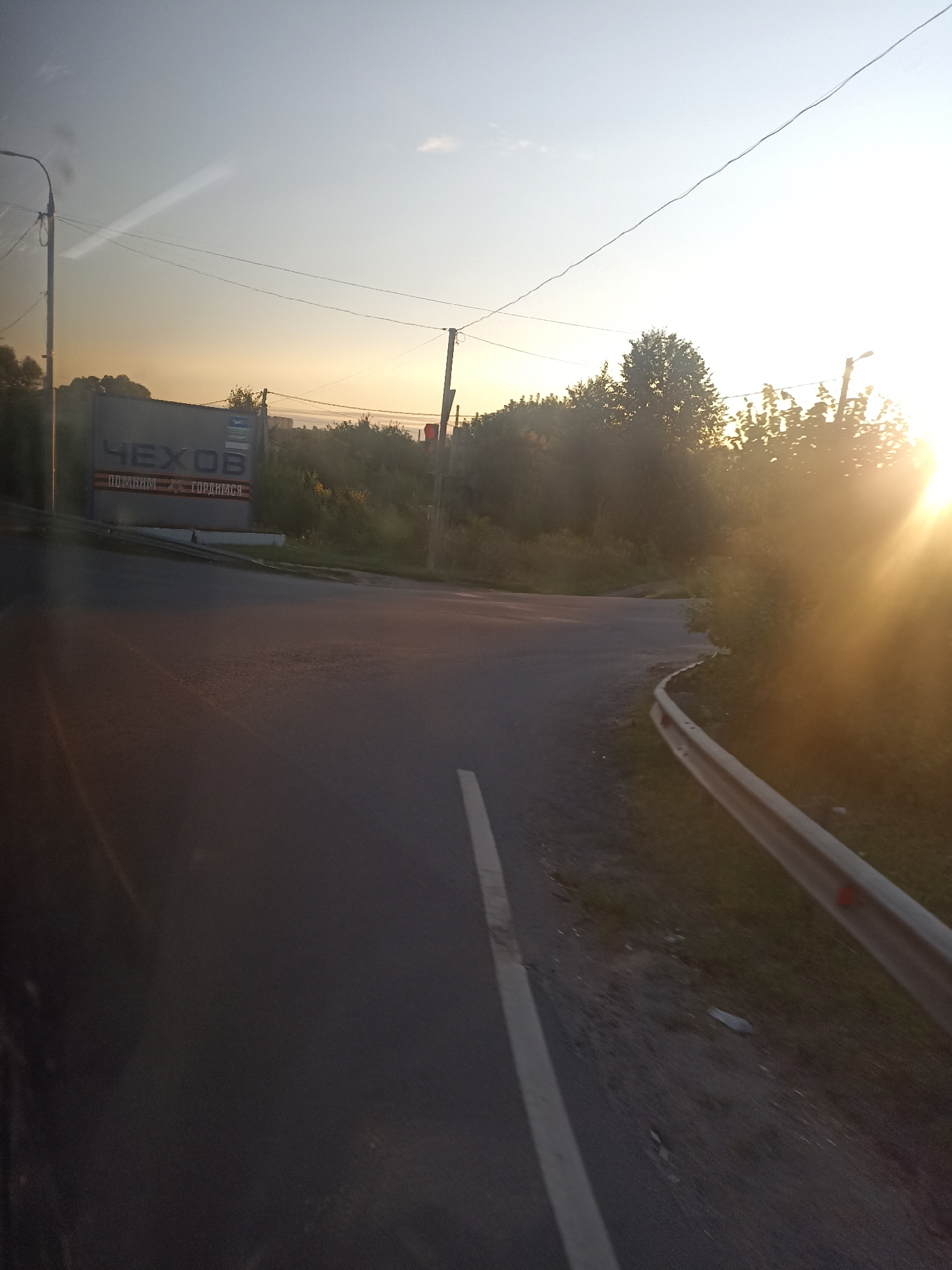 The image depicts a road junction in the city of Chekhov, Russia, during sunset. The sky is a gradient of pale blue to warm orange, indicating the sun is low on the horizon. On the left side of the road, a large sign displays the word "ЧЕХОВ" in bold letters, with smaller text below that reads "ПОМНИМ - ГОРДИМСЯ" (We remember - we are proud). Adjacent to the sign is a traffic light, which is red. Trees and vegetation line the sides of the road and the surrounding area. A metal guardrail is visible along the right edge of the road. The road surface appears to be asphalt, with a white line marking the lane. The scene is characterized by the warm, hazy light of the setting sun, creating lens flares and illuminating the foliage. There are no people or vehicles clearly visible in the frame.