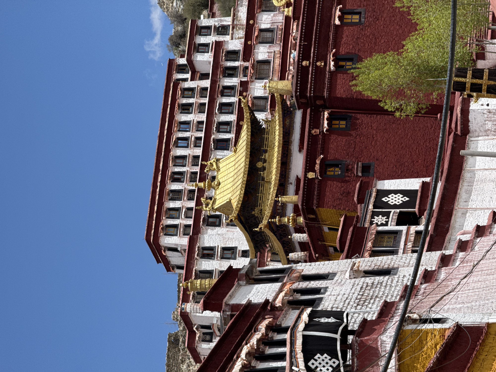 The image captures the Potala Palace in Lhasa, China. The palace, a multi-storied structure, features a predominantly red and white color scheme, with golden architectural elements. The building has numerous windows. Prayer flags are visible near the lower portion of the palace. The sky is blue. A portion of a mountain is visible near the palace. A tree is near the palace.