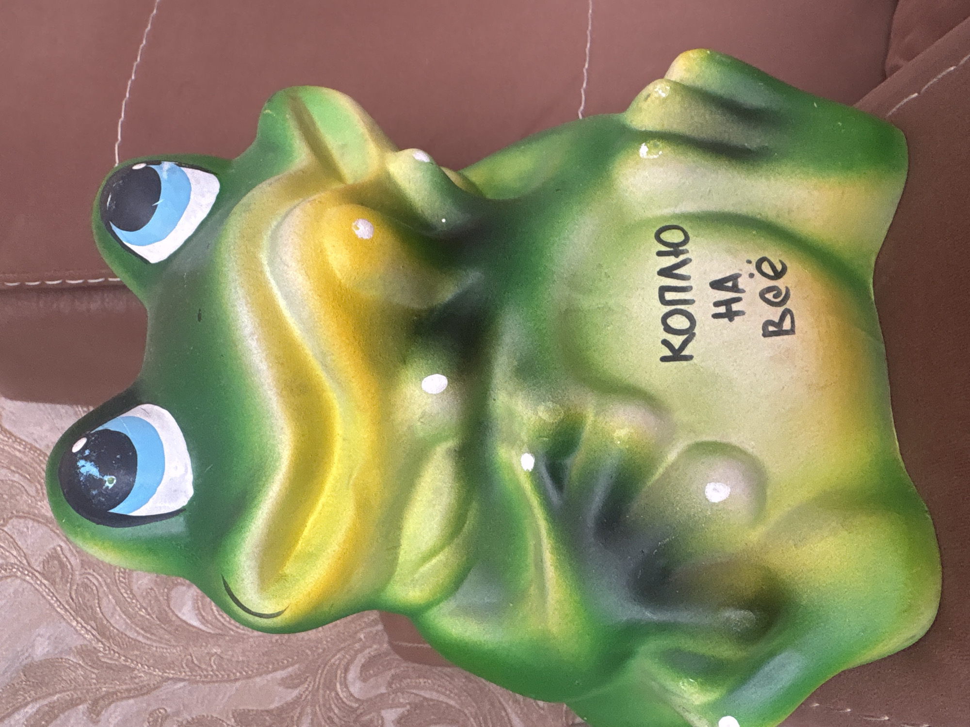 The image displays a green and yellow frog-shaped figurine, likely a piggy bank, resting on a brown upholstered surface. The frog is positioned with its head towards the upper left. Its body features a gradient of colors, primarily green, transitioning to a lighter yellow-green on its belly and some raised contours. The frog has large, prominent blue eyes with black pupils and white highlights, giving it an alert or cheerful expression. A small, curved black line forms a smile beneath its left eye. Minor wear is visible, particularly as chipped paint around the eyes. Several small white dots are painted on its back and sides, possibly as decorative elements.
Prominently written in black Cyrillic script on the frog's back are the words "КОПЛЮ НА ВСЁ" (KOPLYU NA VSYO), which translates to "I am saving for everything." This inscription strongly indicates its function as a money box.
The background consists of a smooth, brown, possibly leather or vinyl, upholstered surface, characteristic of a sofa or chair. A section of patterned fabric, featuring cream and brown swirling designs, is visible in the bottom left corner, beneath the frog. The lighting is soft and diffused, consistent with an indoor environment. The image captures a static object, with no discernible activity taking place. The item is located in Krasnoyarsk, Russia.