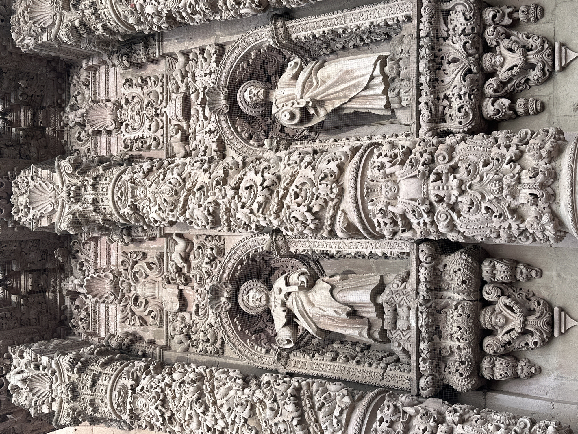 The image shows a detailed view of the highly ornate facade of the Zacatecas Cathedral in Zacatecas City, Mexico. The facade is composed of intricate stone carvings, featuring columns, statues, and decorative motifs. Two prominent statues of male figures, likely saints, are visible within arched niches. They are dressed in robes and hold symbolic objects, though their identities are not clearly discernible from this angle. The stonework is heavily detailed, with layers of relief carvings depicting cherubs, floral elements, and geometric patterns. Fine mesh netting is visible in front of the statues and some of the decorative elements, likely to protect them from the elements or birds. The overall impression is one of elaborate Baroque architecture, characterized by its richness and complexity.