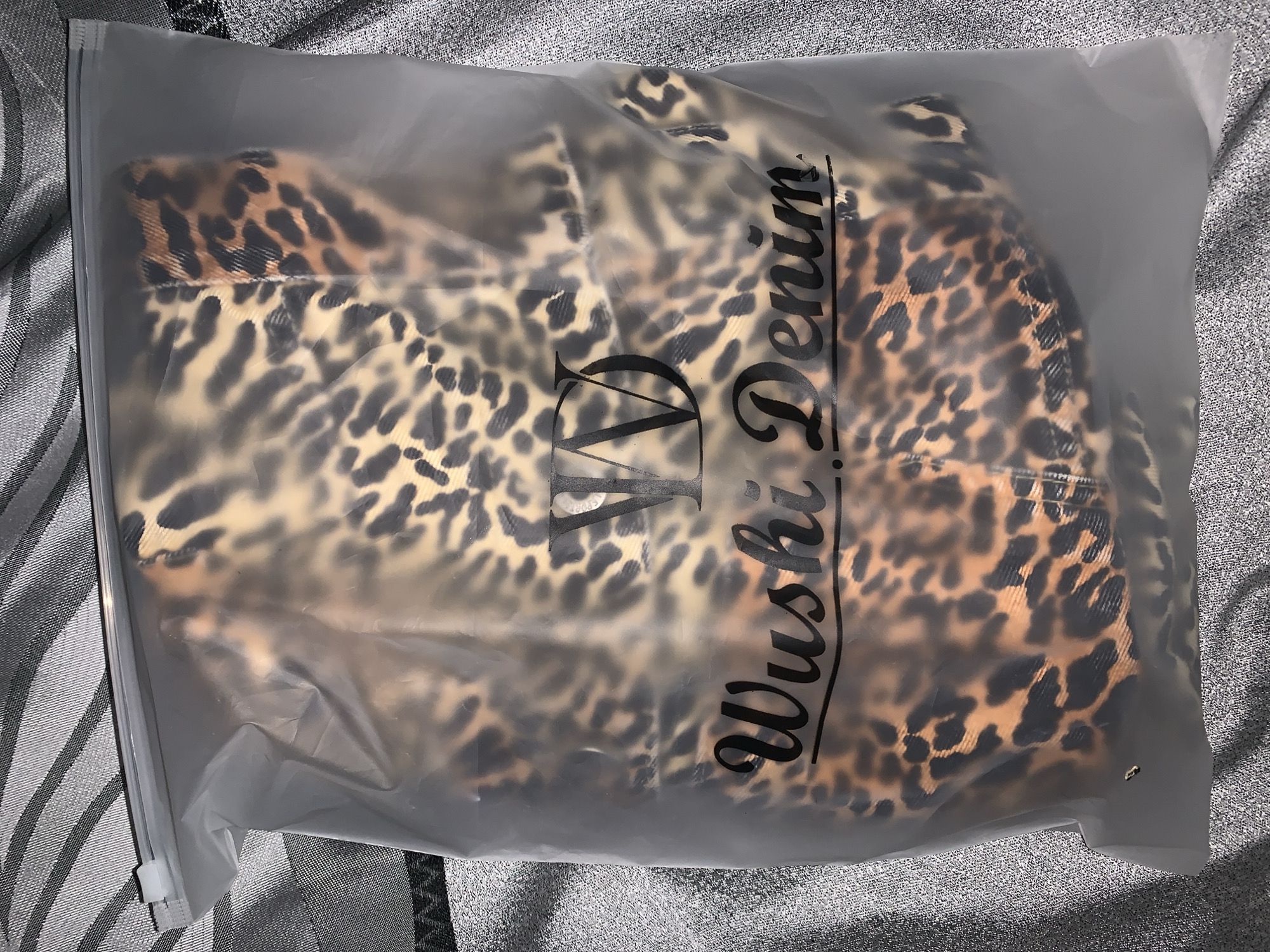 A semi-transparent plastic bag containing a folded garment with a leopard print is placed on a silver-gray textured surface. The bag features black text that reads "Wushi Denim" vertically. The scene is indoors and there is no visible evidence to indicate this is located in the city of Rafin Par, Nigeria.