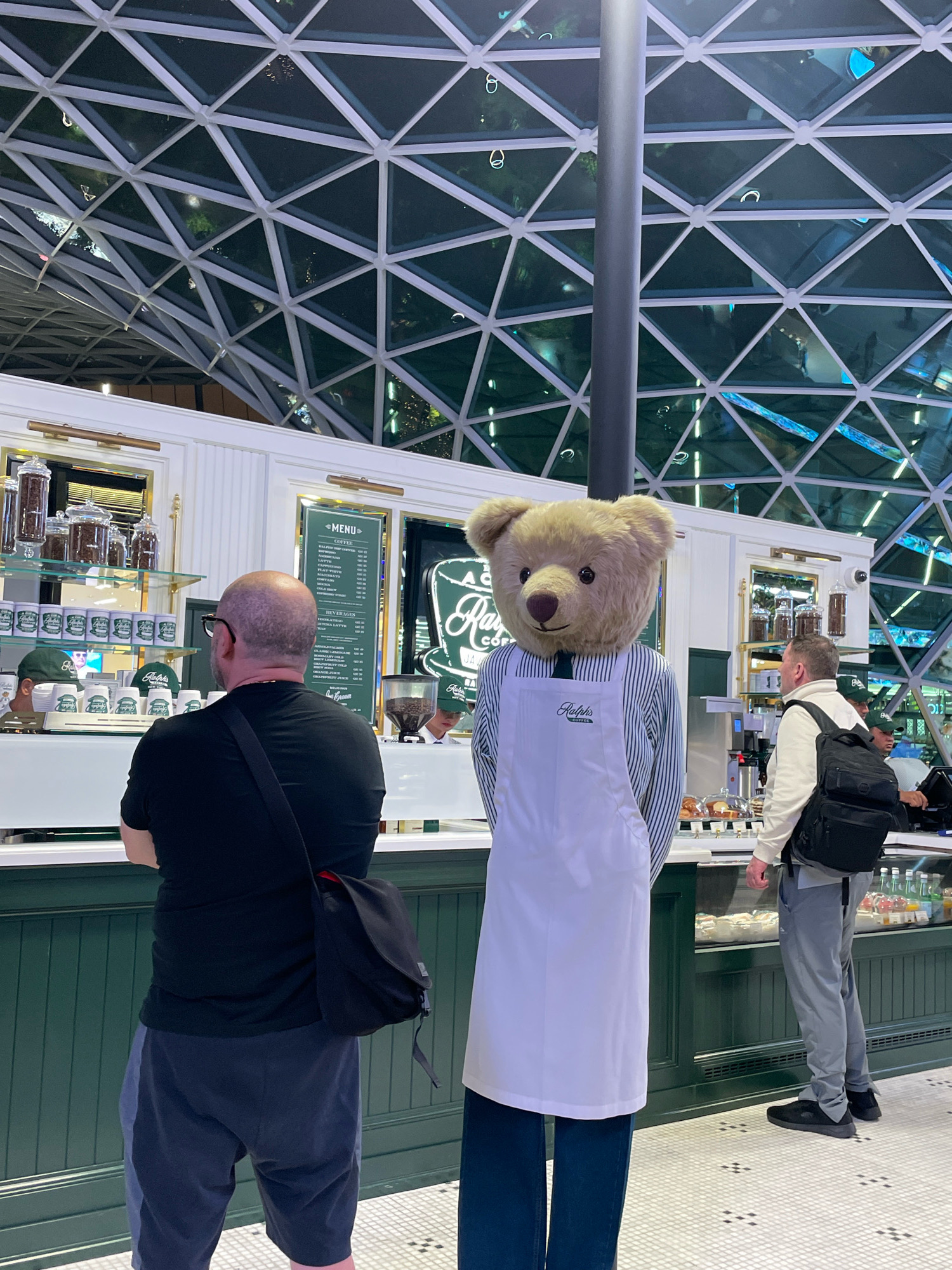 In Doha, Qatar, at a Ralph's Coffee establishment, a person in a teddy bear mascot costume, wearing a striped shirt and white apron, stands facing away from the camera. To the left of the mascot, a bald man wearing glasses and a black t-shirt with a messenger bag slung across his body is facing the counter. To the right of the mascot, a man with a black backpack is seen from the side, also facing the counter. The background features a brightly lit coffee shop interior with a glass ceiling structure overhead, showcasing a geometric pattern of triangles. The counter displays food items and coffee-making equipment.
