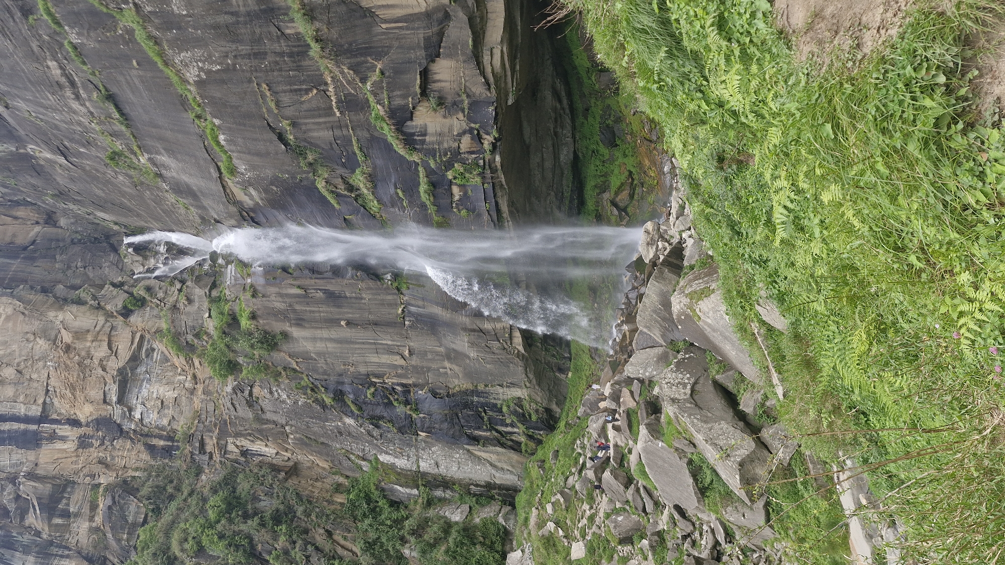 A tall, multi-tiered waterfall cascades down a sheer, rocky cliff face. The water is a bright white, creating a misty spray at the bottom. The cliff is composed of layered rock, with dark streaks and patches of green vegetation clinging to its surface. Below the waterfall, a jumble of large, grey rocks lies scattered, some partially covered by lush green grass and ferns. Several people, small in scale compared to the immense natural feature, are visible standing among the rocks near the base of the waterfall. They appear to be observing the scene. The environment is natural and rugged, with a sense of grandeur and untouched beauty. The location is identified as the city of Bahang, India.
