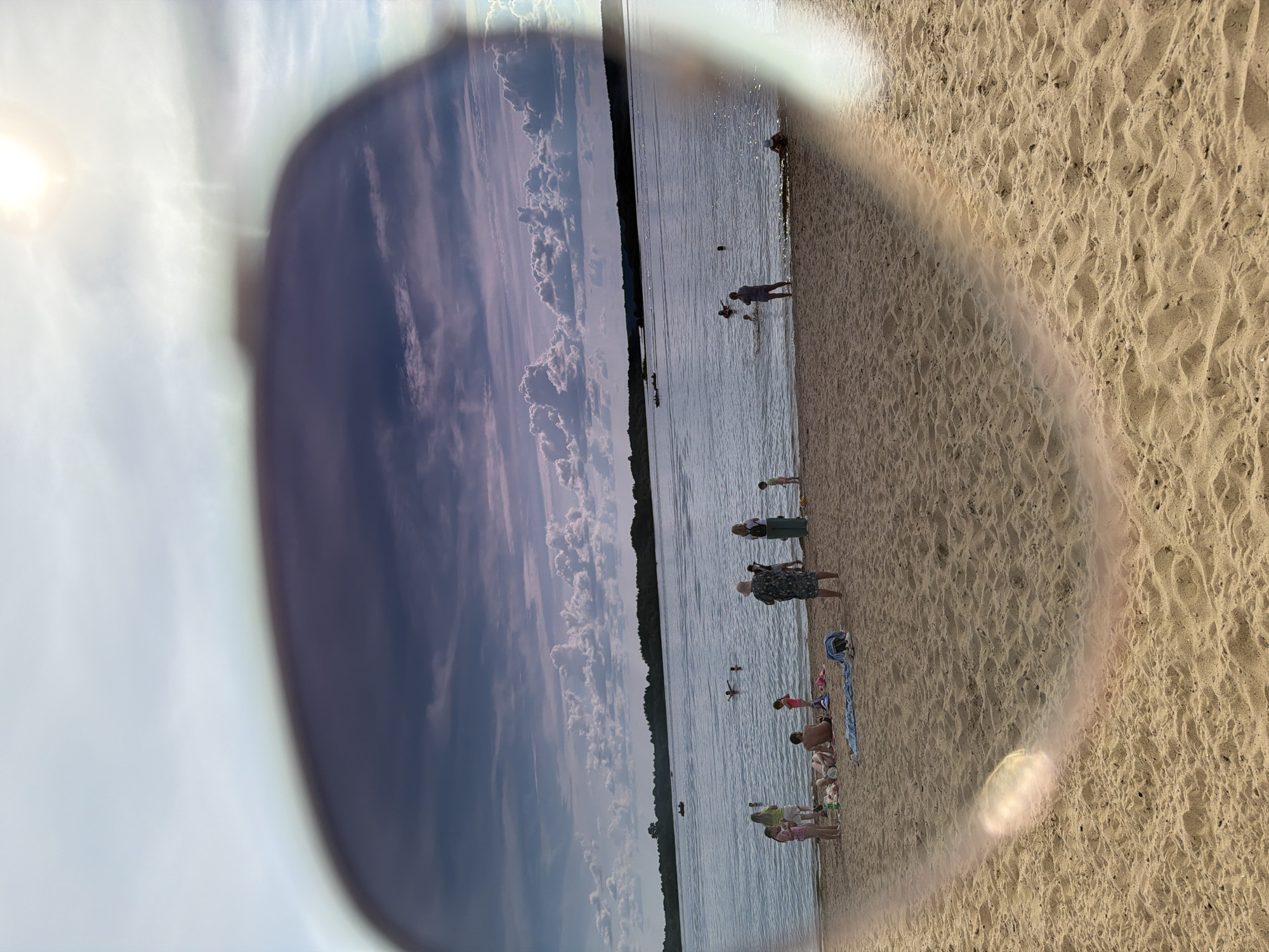 The image depicts a beach scene on a partly cloudy day. The foreground is dominated by sand, with footprints and slight ripples indicating recent activity. In the mid-ground, a body of water, likely a river or lake, is visible, with several people swimming and wading. Along the shoreline, there are groups of people sunbathing and standing, some appear to be families. Boats are visible on the water in the distance, and a tree line borders the far side of the water. The upper portion of the image shows a sky with a mix of blue sky and dramatic clouds, with the sun appearing bright and high in the sky, casting a glare that partially obscures parts of the image. The overall impression is of a recreational setting. This image does not contain any identifiable landmarks or features that would place it specifically in the city of Solikamsk, Russia.