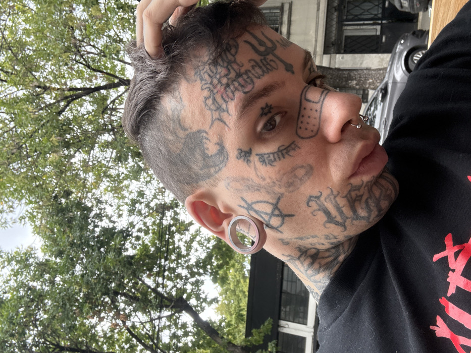 A heavily tattooed individual is depicted in a close-up, outdoors on an urban street in Buenos Aires, Argentina. The subject, appearing male, is positioned slightly off-center, looking downward and to the viewer's left. They have dark hair, styled short on the sides with a longer, textured top, and are shown raising their left hand (viewer's right) to touch their hair. Numerous black ink tattoos cover their entire visible face, neck, and scalp, including script, symbols such as an anarchy sign and a trident, and a stylized band-aid under the left eye. A septum piercing with a silver captive bead ring is present on the nose, and a large silver-colored tunnel earring is visible in the stretched left earlobe. The individual wears a black long-sleeved shirt with red graphic text partially visible on the chest.
The background shows an overcast sky and abundant green foliage from trees, suggesting a tree-lined street. In the mid-ground, a dark-colored building or structure and a light grey sedan car are partially visible, parked on what appears to be a paved street or sidewalk. The scene is illuminated by soft, diffuse light consistent with an overcast day.