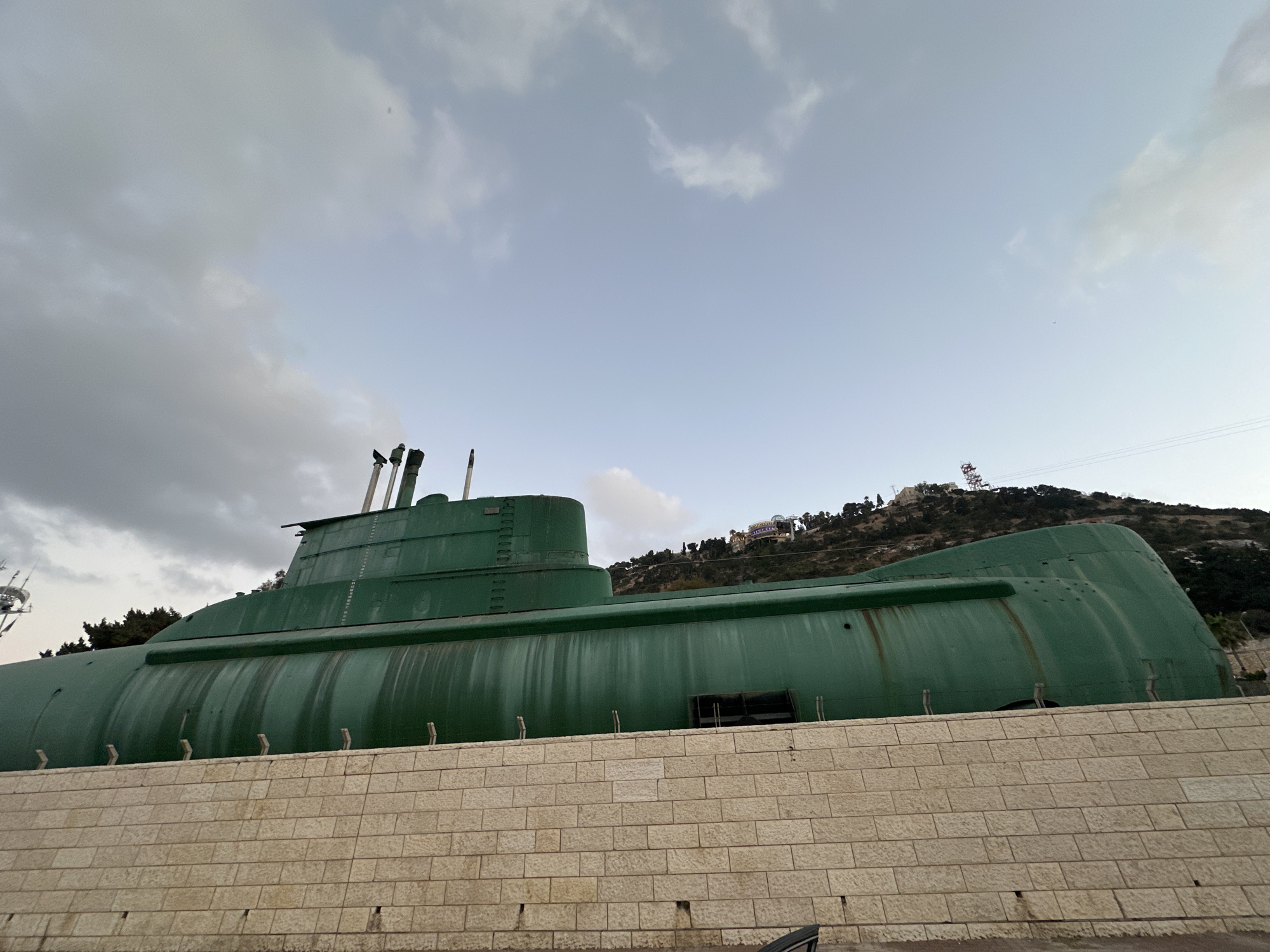 An exterior, low-angle, wide shot captures the side of a large, weathered green submarine resting on land in Haifa, Israel. The submarine is positioned behind a light-colored, rough-hewn stone wall that runs horizontally across the lower portion of the frame. The submarine's hull displays streaks of rust and grime, indicating age and exposure to the elements. Several small, vertical metal posts protrude from the top of the stone wall, directly in front of the submarine.
The submarine's conning tower, also painted green, is visible towards the left side of the image, with several cylindrical structures, likely periscopes or antennas, extending upwards. The background consists of a steep, tree-covered hillside under a pale, overcast sky with scattered clouds. Structures, possibly buildings or a tower, are visible on the hilltop. The overall scene conveys a sense of historical preservation or a static display.