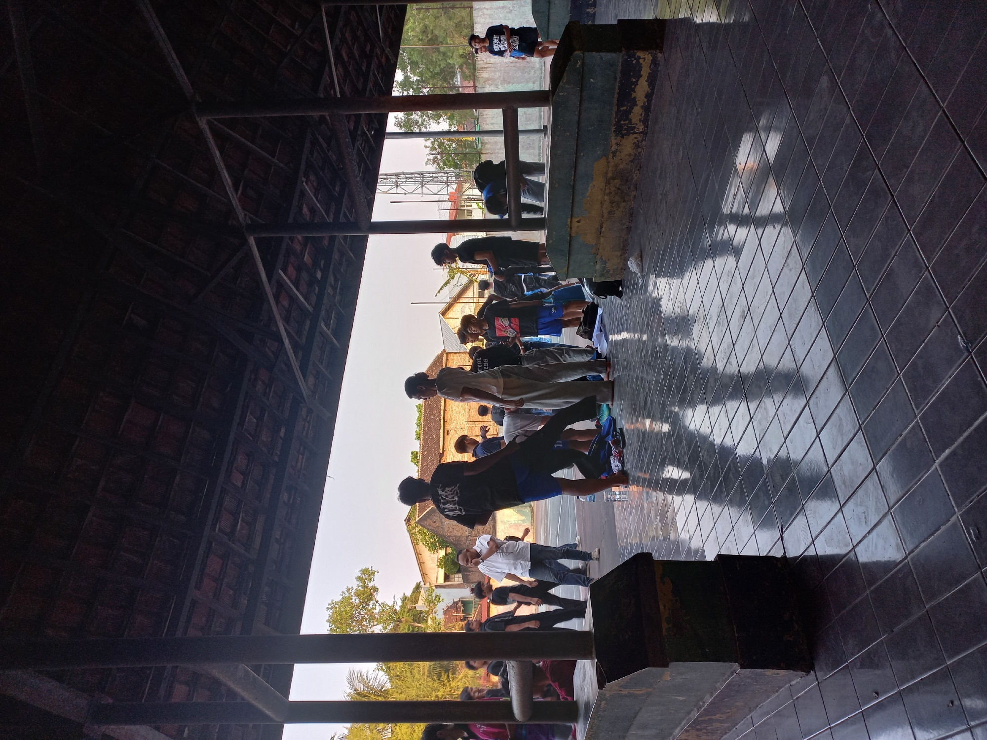 The image depicts an outdoor gathering beneath a wooden-tiled roof supported by pillars. The ground is paved with dark, glossy tiles that reflect the overhead structure and the people present. In the foreground, two large, dark, rectangular structures with weathered yellow trim are visible. A group of young men and a few adults are dispersed throughout the scene, engaged in various activities.
Some individuals are standing and conversing, while others appear to be preparing for or concluding an activity. One man in a white shirt and jeans is standing with his hand to his face, appearing to be speaking on a phone. Another man in a black t-shirt with graphics is leaning over. In the background, buildings with red-tiled roofs and trees are visible under a bright sky. The overall impression is a casual gathering in a semi-public outdoor space, possibly a community center or a sports complex, given the presence of what looks like a sports court in the background. The atmosphere suggests a warm day with the sun casting strong reflections on the tiled floor.