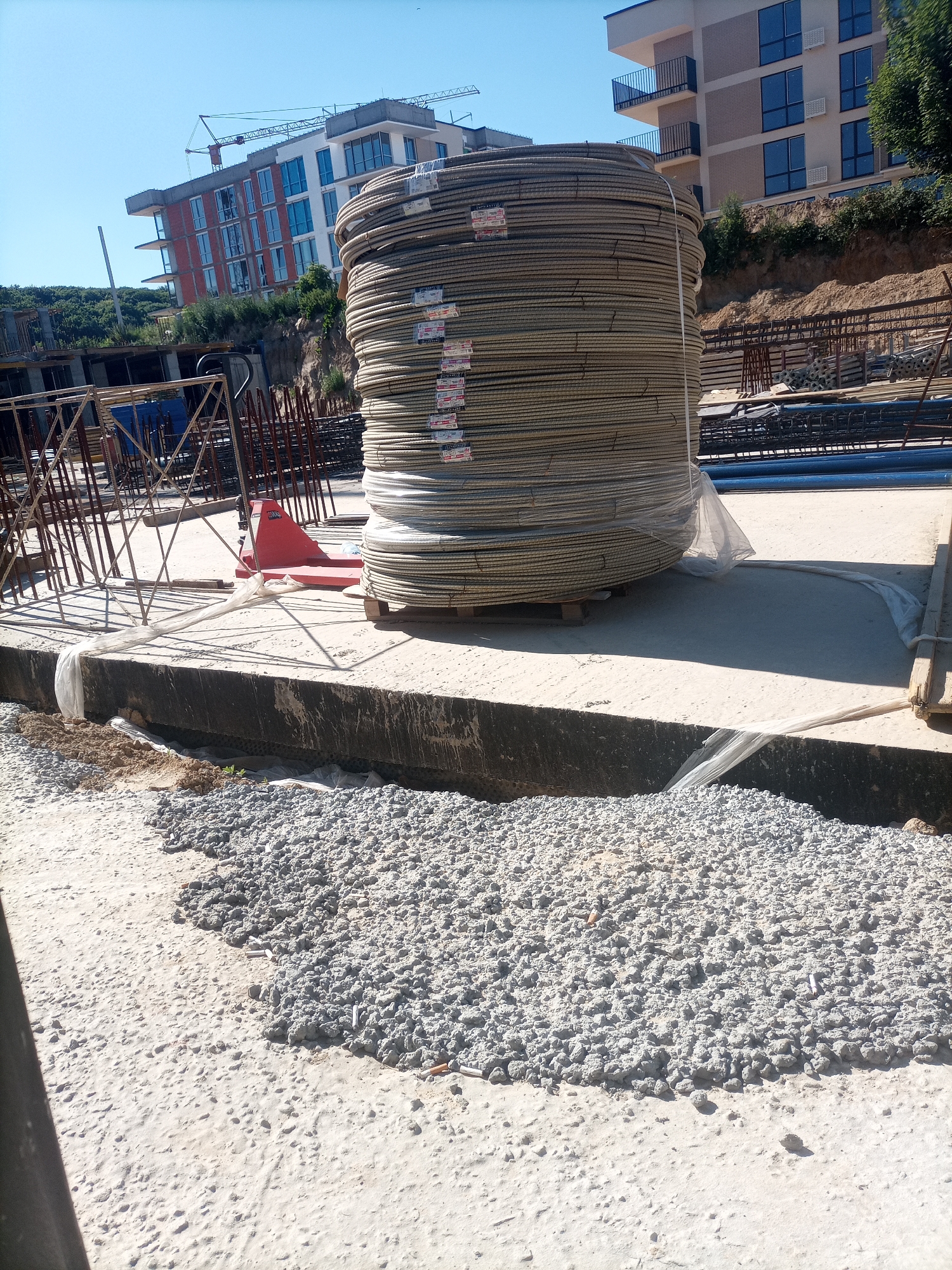The image depicts a construction site under a clear blue sky. In the foreground, a large pile of coiled rebar, wrapped in clear plastic, dominates the scene. The rebar is stacked on a wooden pallet, with a red pallet jack visible to its left. Adjacent to the rebar, a concrete foundation runs horizontally across the frame, partially covered with light-colored plastic sheeting. A trench filled with gravel and some dirt runs along the front edge of the foundation. In the background, two multi-story buildings are under construction. One building, closer to the viewer, has a red brick facade and multiple windows. Another building, further back, has a light-colored facade with balconies and windows. Construction cranes are visible above the buildings. The overall environment suggests an active construction zone.