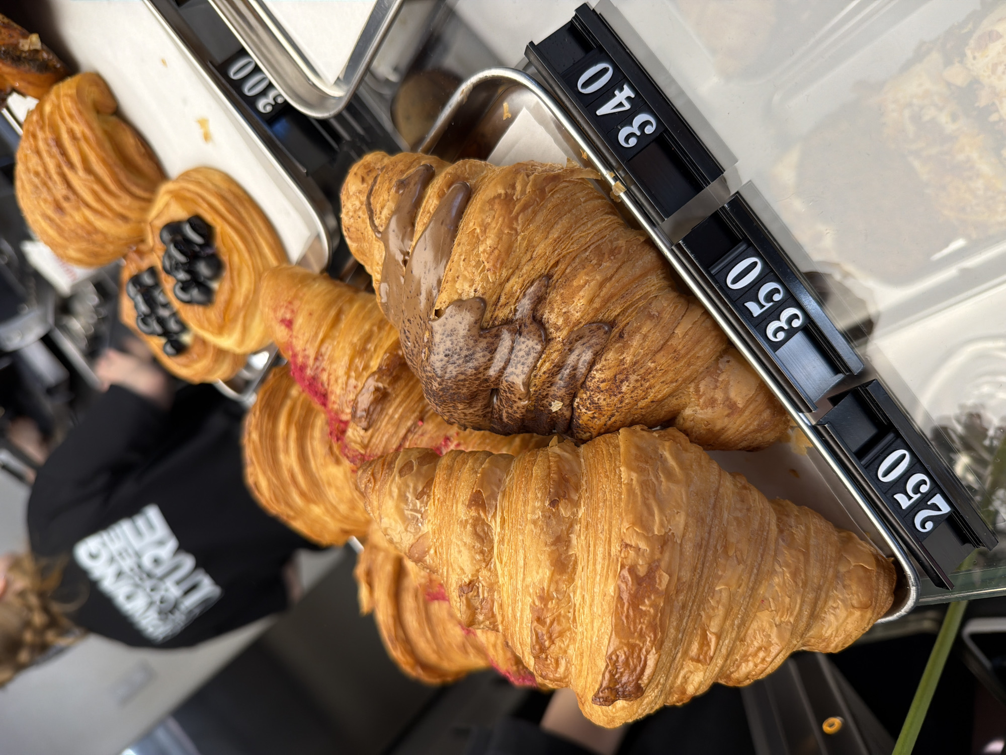 The image shows a display of baked goods in a cafe or bakery. The primary subjects are croissants and other pastries, with prices displayed next to them. A sign indicates a chocolate-covered croissant costs 340 (currency unknown), a plain croissant costs 250 (currency unknown). The pastries are arranged on metal trays. Part of a person is visible on the left, wearing a black garment. Based on the image alone, there is no information that allows us to identify the location is in Izhevsk, Russia.