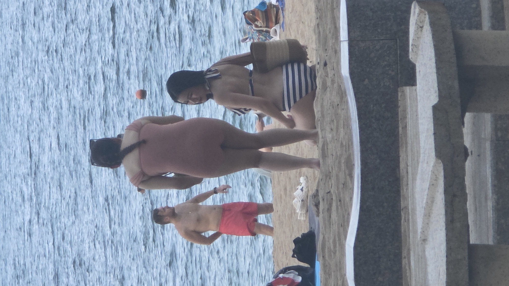 This daytime image depicts a beach scene in Nha Trang, Vietnam, with people enjoying the calm waters and sandy shore. The photograph is rotated 90 degrees clockwise.
In the foreground, standing in the shallow, calm blue water, is a woman wearing a light pink one-piece swimsuit. Her dark hair is pulled back, and she has sunglasses on her head, facing away from the camera. Beside her, closer to the viewer and also in the water, is a shirtless man with a beard, wearing red swim shorts. He is partially turned, looking towards the water. A small orange buoy floats in the distance on the water's surface.
Further back on the sandy beach, another woman is visible, also facing away. She is wearing a black and white striped bikini and is holding a woven straw bag over her shoulder. Behind her, a colorful striped beach bag and other items are scattered on the sand. The sand itself is light-colored and forms a gentle slope towards the water. On the far right, a sturdy, multi-tiered concrete or stone structure frames the scene, suggesting a promenade or wall adjacent to the beach.
The overall atmosphere is one of leisure and relaxation on a clear, sunny day at the beach. No visible text is present in the image.
