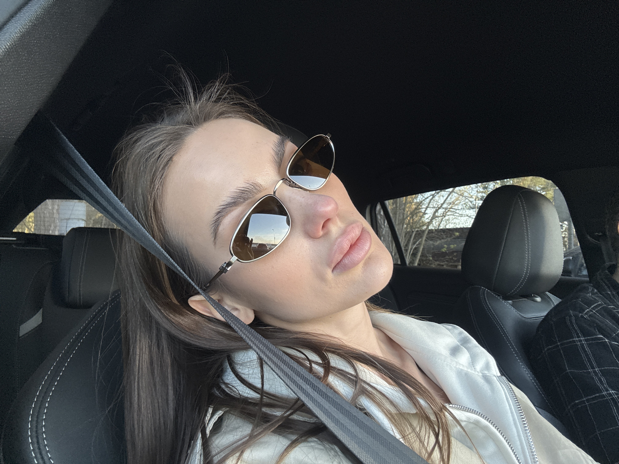 A young woman is captured in a close-up, chest-up shot inside a car, positioned as a passenger. Her head is tilted back and to the left, revealing her profile. She wears gold-rimmed oval sunglasses that reflect a bright, clear sky and distant trees. Her lips are pursed in a "duck face" expression. Her long, dark brown hair flows over her shoulders and down her back. She is dressed in a light-colored, possibly cream or white, zip-up jacket. A dark-colored seatbelt with a textured pattern crosses diagonally over her chest. The car's interior is dark, featuring black leather seats with white stitching. Outside the window, bare trees are visible under what appears to be a clear, sunny sky, indicating daytime. A second person, wearing a black and white plaid garment, is partially visible in the adjacent seat. The image conveys a casual, relaxed moment. No visible text is present in the image. The setting is Białobrzegi, Poland.