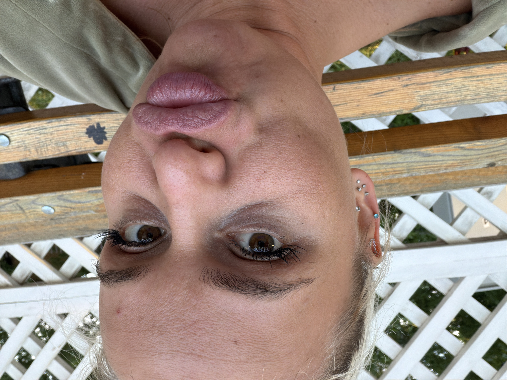 A close-up, inverted selfie depicts a person with light skin, light eyebrows, and blonde hair. The person has brown eyes, with eyelashes visible and some eyeliner. Their lips are pursed. They are wearing multiple earrings in their left ear. The background consists of a light-colored wooden bench with a four-leaf clover design and a white lattice structure. The location is most likely outside due to the presence of greenery visible through the lattice. There is no specific indication that the location is in Sarapul, Russia.