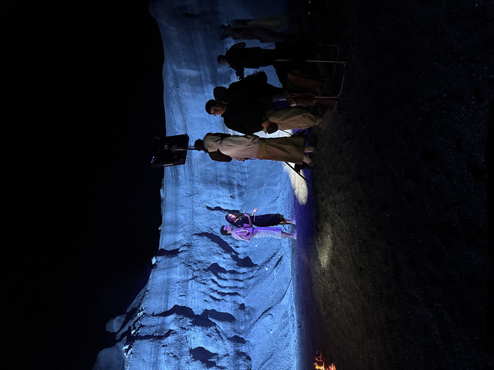 The image shows a scene being filmed at night in a cave. Two individuals are lying on a rock surface, illuminated by blue light. A film crew, including people sitting on chairs and operating equipment, is present near the top of the frame. A light with the brand "Amaran" on it is visible. Flames from a fire are at the bottom of the frame. This image was taken in the city of Teguise, Spain.