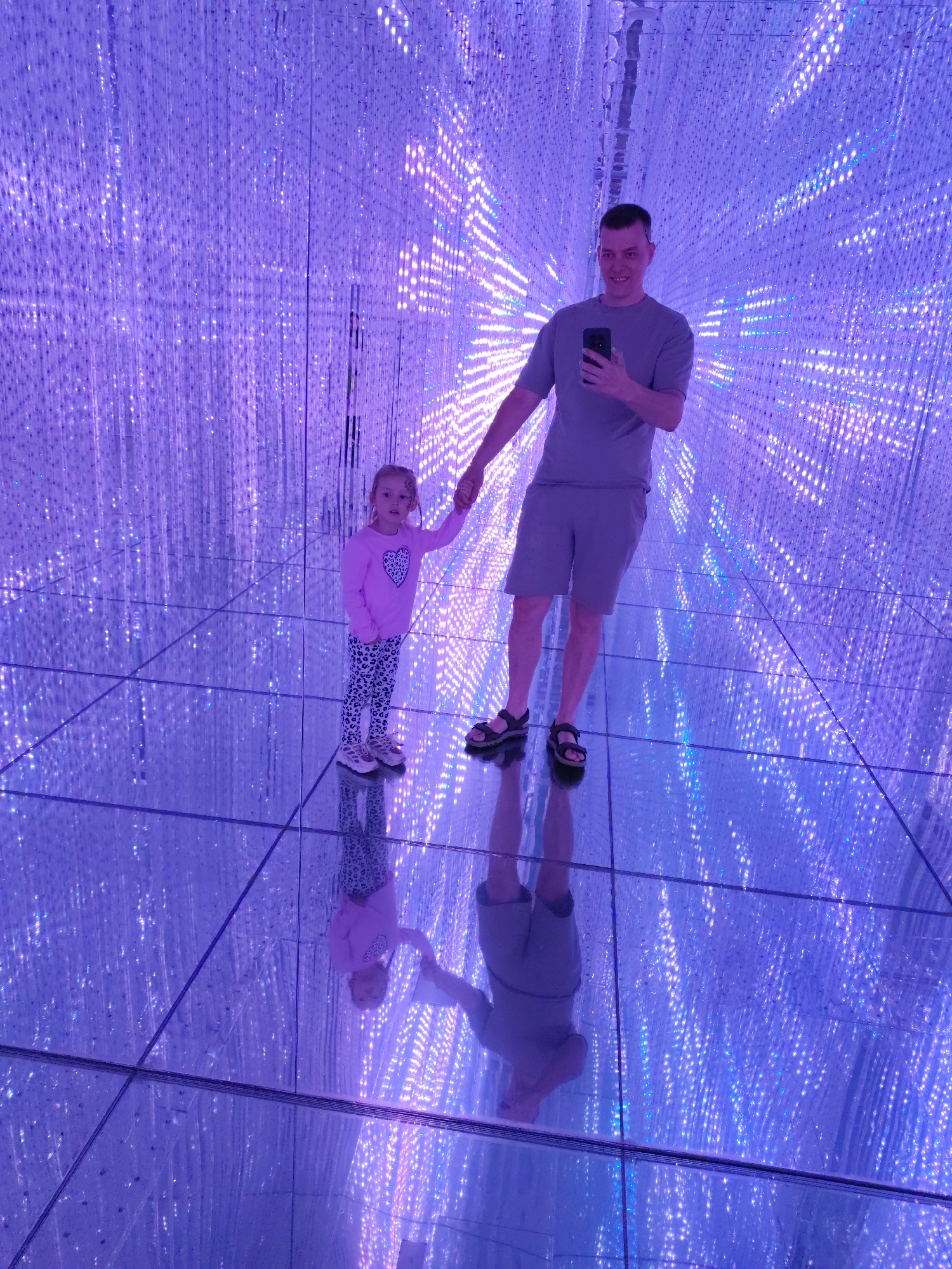 An indoor scene in Kazan, Russia, depicts a man and a young child holding hands in a room with reflective walls, ceiling, and floor. Both are reflected on the floor. The walls and ceiling are adorned with many vertical light strands that create a tunnel-like effect. The man wears a gray t-shirt and shorts with sandals. The child has a pink long-sleeved shirt with heart design, leopard-printed leggings, and sneakers. The man is holding a mobile phone in his left hand, pointed towards himself as if taking a selfie.